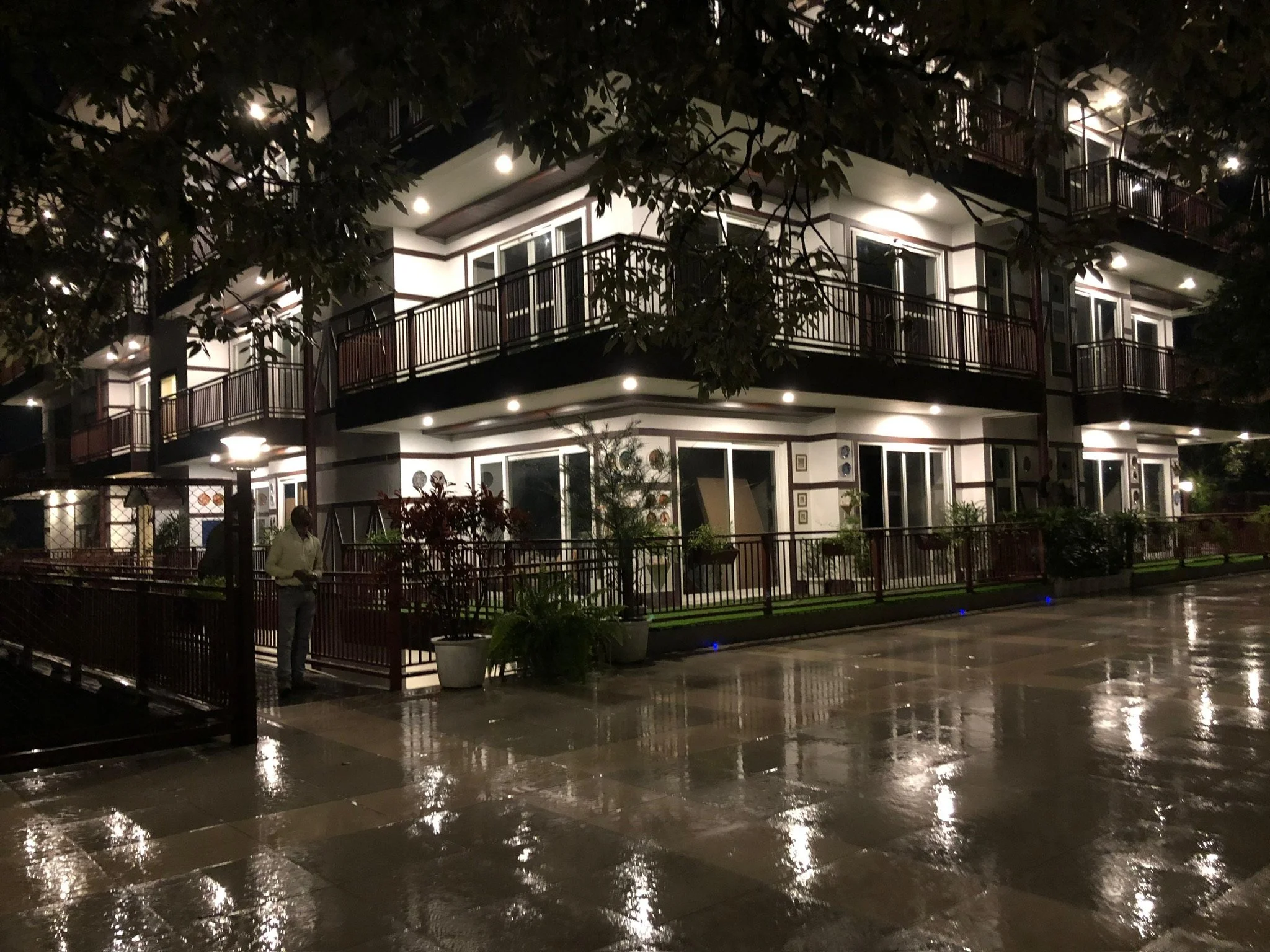 Nighttime scene of a multi-story apartment building with lit exterior lights, a wet reflective paved walkway, and a person standing near the fence.