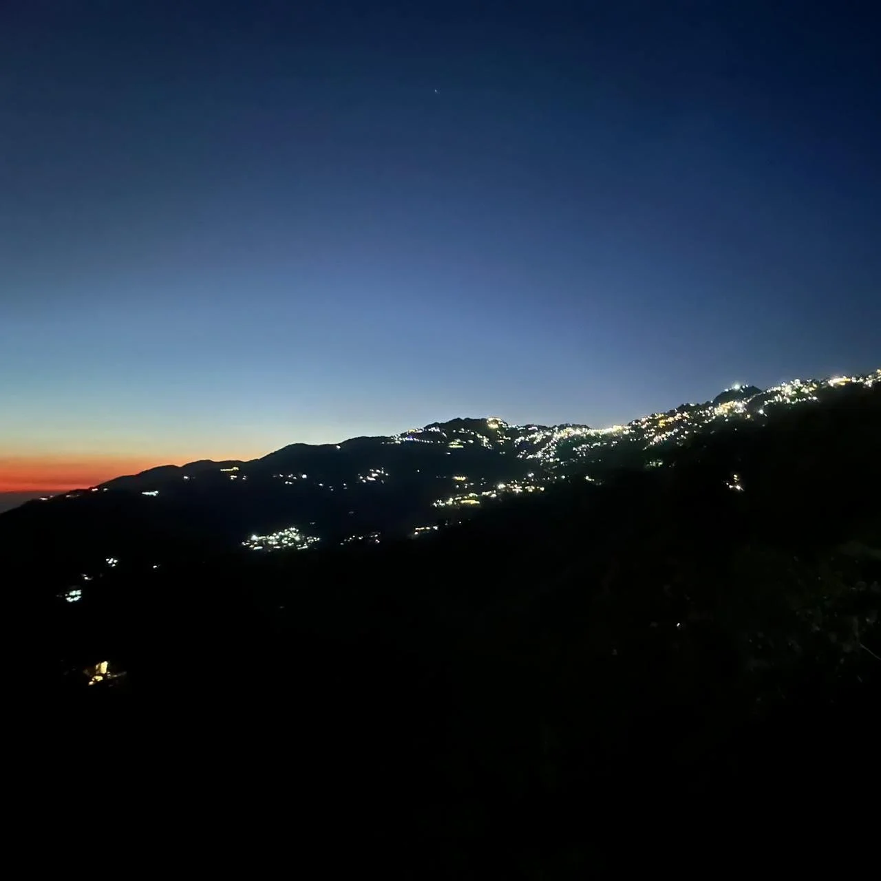 Nighttime landscape of a mountain range with scattered lights from houses, under a darkening sky with a faint sunset on the horizon.