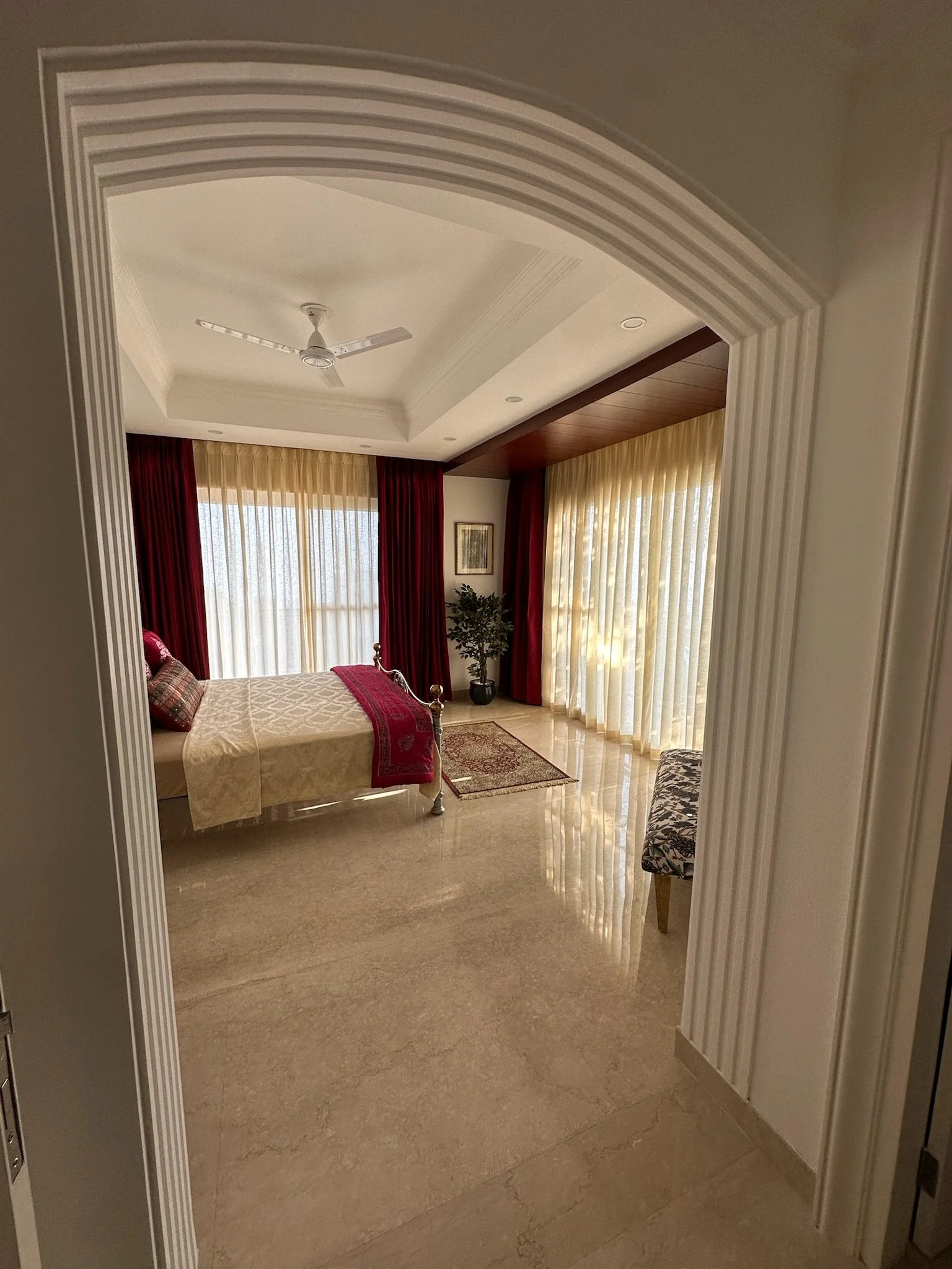A bedroom with a bed, red and cream curtains, a framed picture, a potted plant, and a wooden floor.
