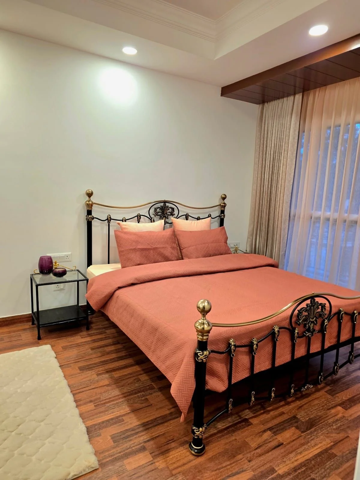 A bedroom with a vintage black and gold metal bed, with a pink bedding set, located near a window with beige curtains, with a small black side table holding purple decorative items and a cream-colored rug on a wooden floor.