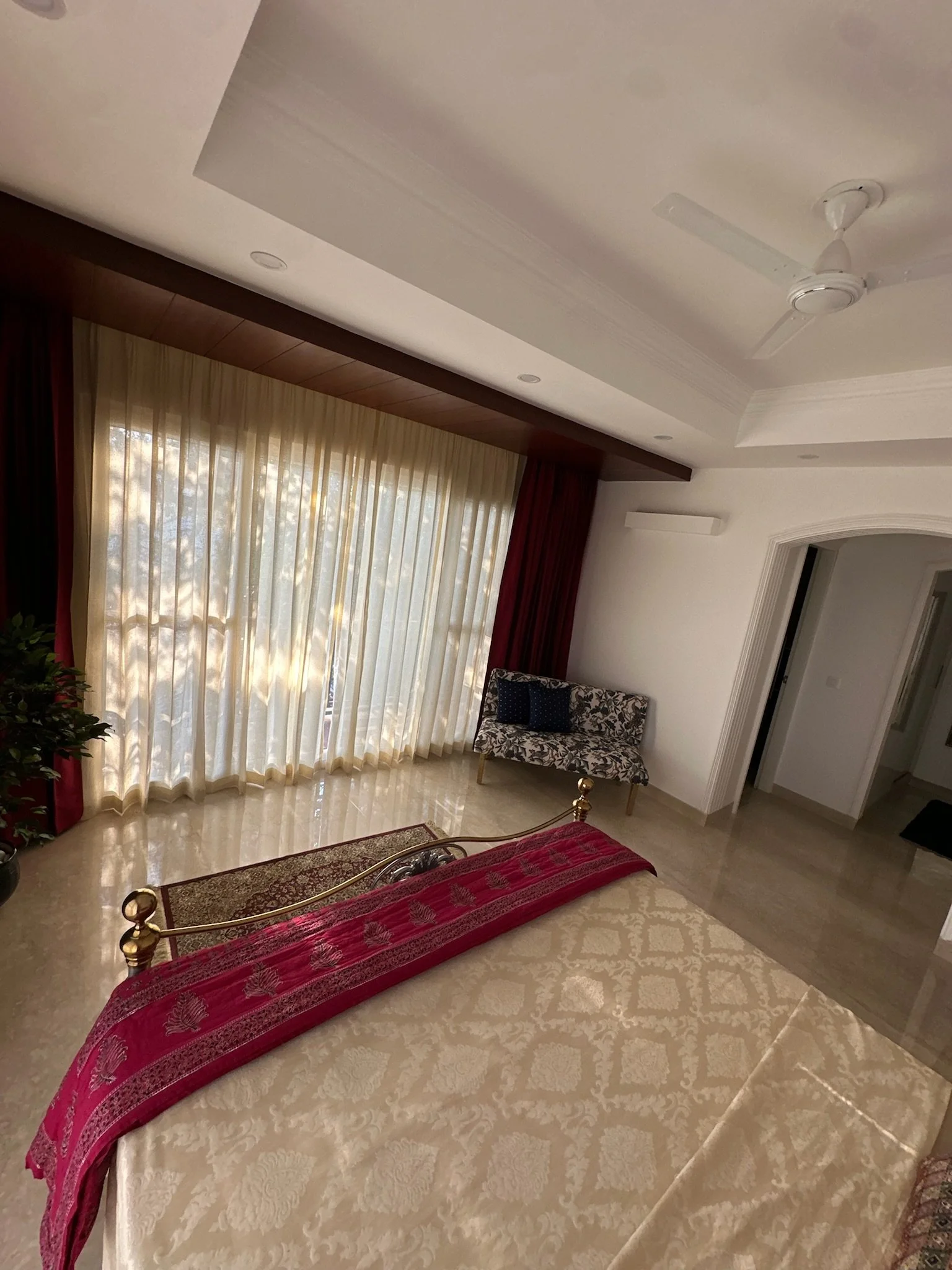 A potted leafy green plant in a black pot with white stones, situated between red and cream curtains, with a framed artwork on a white wall above.