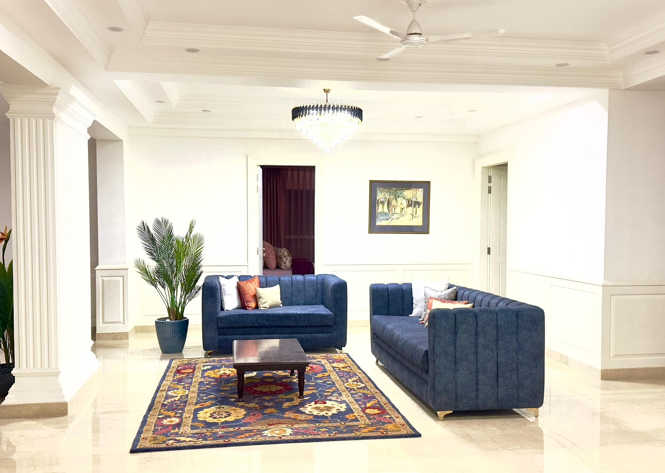 A spacious living room with white walls, a ceiling fan, a chandelier, two blue sofas with pillows, a wooden coffee table, a patterned rug, a tall green potted plant, and a framed painting on the wall.