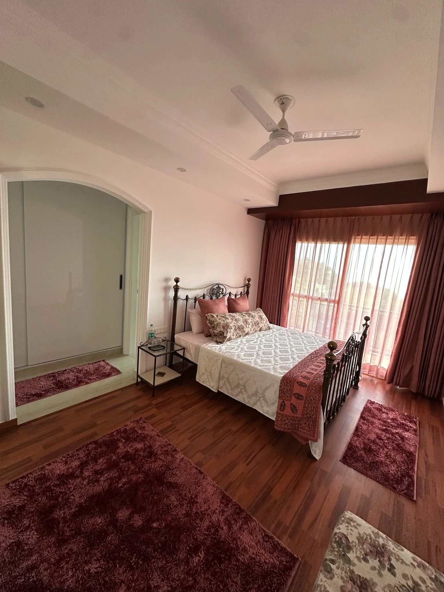 A bedroom with a black and gold ornate metal bed frame, white bedding with a floral pattern, blue pillows, and a padded turquoise chair next to a small black desk with a pink flower arrangement.
