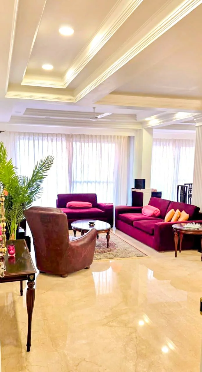 Living room with beige marble floors, white crown molding ceiling, purple sofas with pink pillows, an armchair, a small round coffee table, a potted plant, and large windows with white curtains.