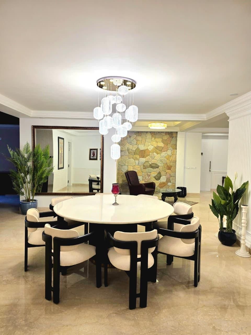 A dining room with a round table, ten chairs, a chandelier, large wall mirror, plants, a stone accent wall, and a small seating area with an armchair.