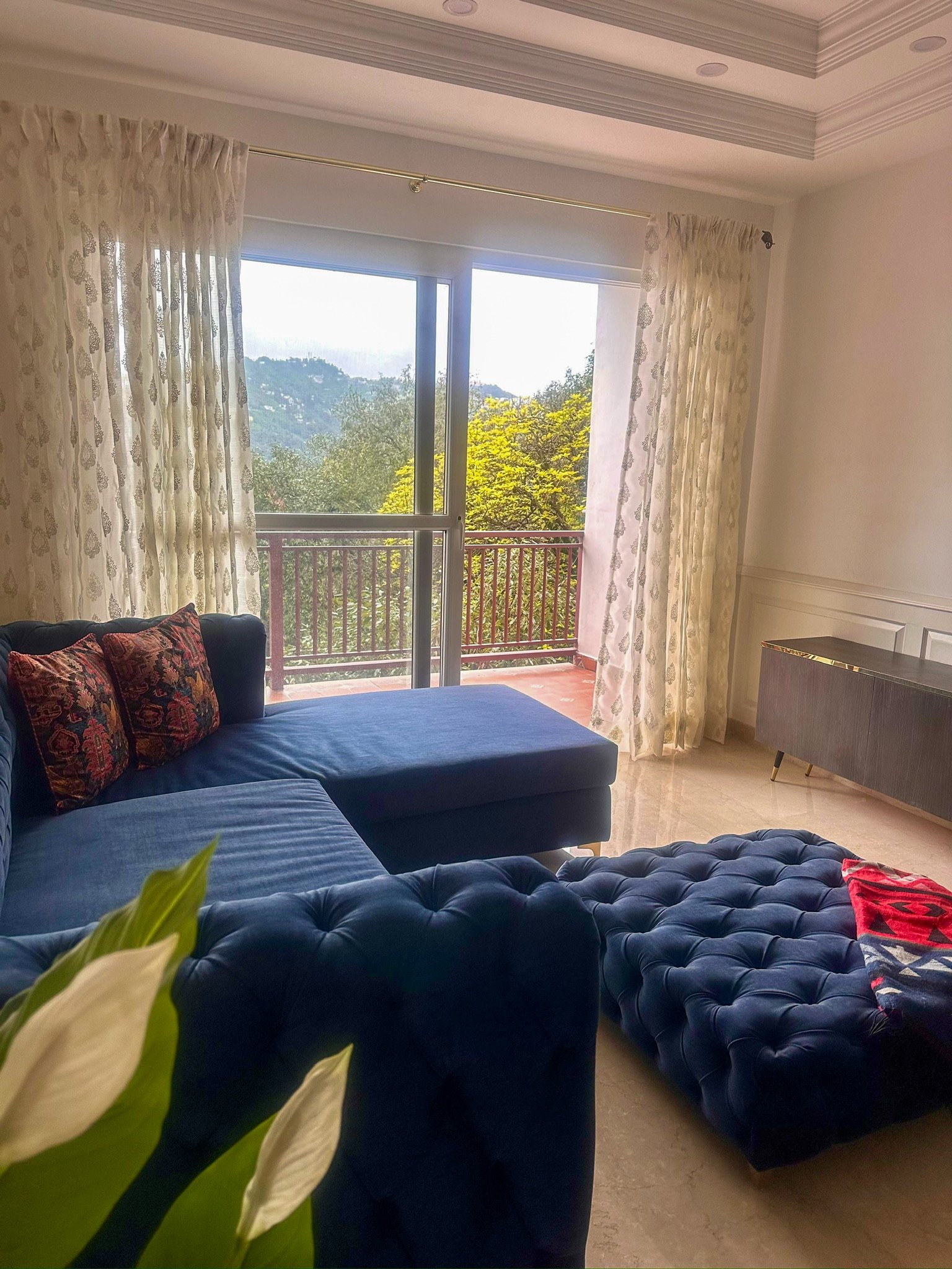 Living room with cream-colored curtains, a sliding glass door leading to a balcony with a view of trees and distant hills, a dark blue tufted sofa, a matching ottoman, and a gray sideboard.
