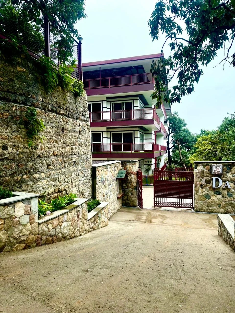 View of a multi-story residential building with balconies, stone walls, and a red gate, surrounded by trees.