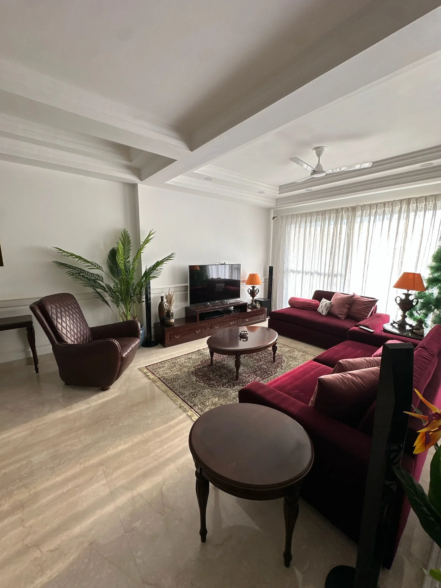 Living room with beige marble floors, white crown molding ceiling, purple sofas with pink pillows, an armchair, a small round coffee table, a potted plant, and large windows with white curtains.