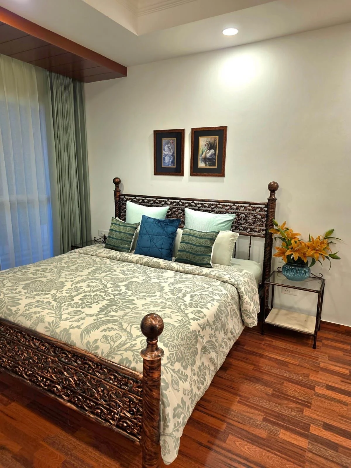 A bedroom with a carved wooden bed, pillows, and a floral bedspread. There are two framed pictures of swans on the wall above the headboard. A side table with a bouquet of yellow lilies is beside the bed, and there are curtains covering a window or sliding door.