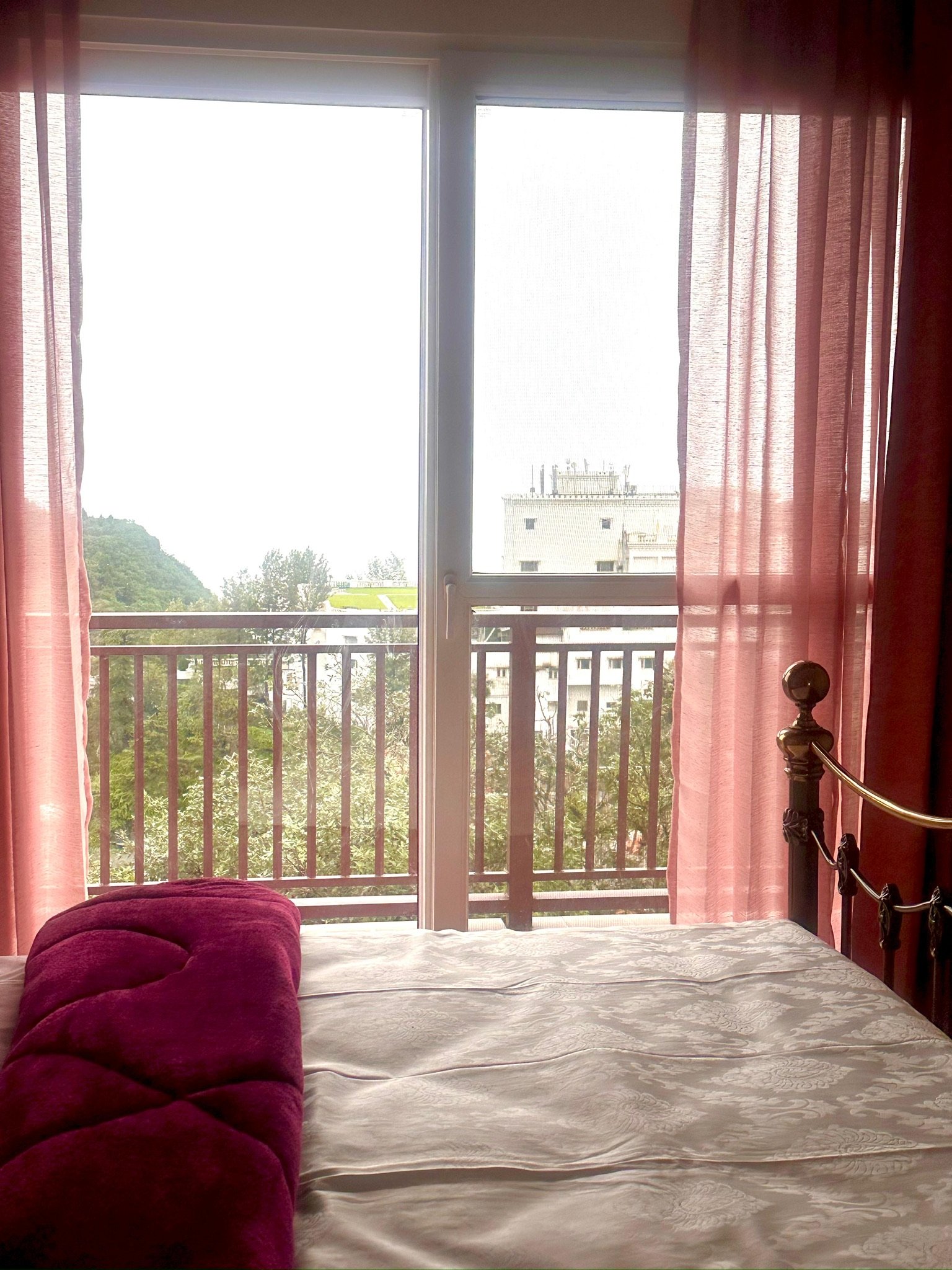 A bedroom with large windows and pink curtains, showing a cityscape and a building in the background. Part of a bed with a decorative pillow and white bedding is visible in the foreground.