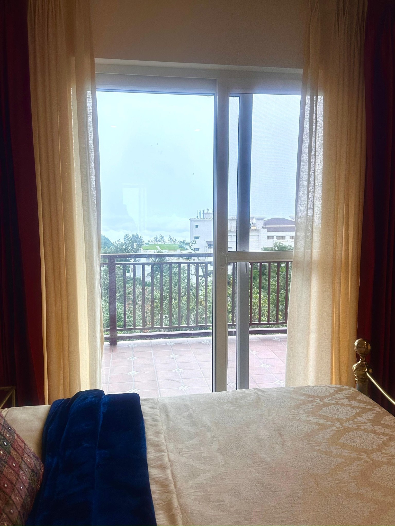 A view of a bedroom with a bed in the foreground, draped in a cream-colored bedspread, and a black and gold bedpost. Behind the bed, there are large glass sliding doors leading to a balcony with a black railing. The balcony has terracotta tiles, and outside, lush green trees and buildings are visible under a partly cloudy sky. Sheer, light-colored curtains frame the doors.