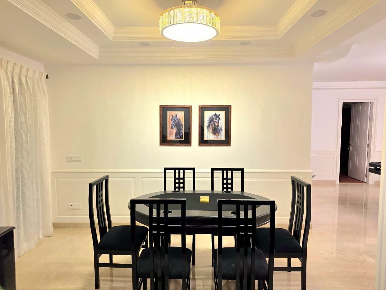 Dining room with six black chairs around a black oval table, two framed horse paintings on the wall, elegant chandelier overhead, and light-colored walls and flooring.