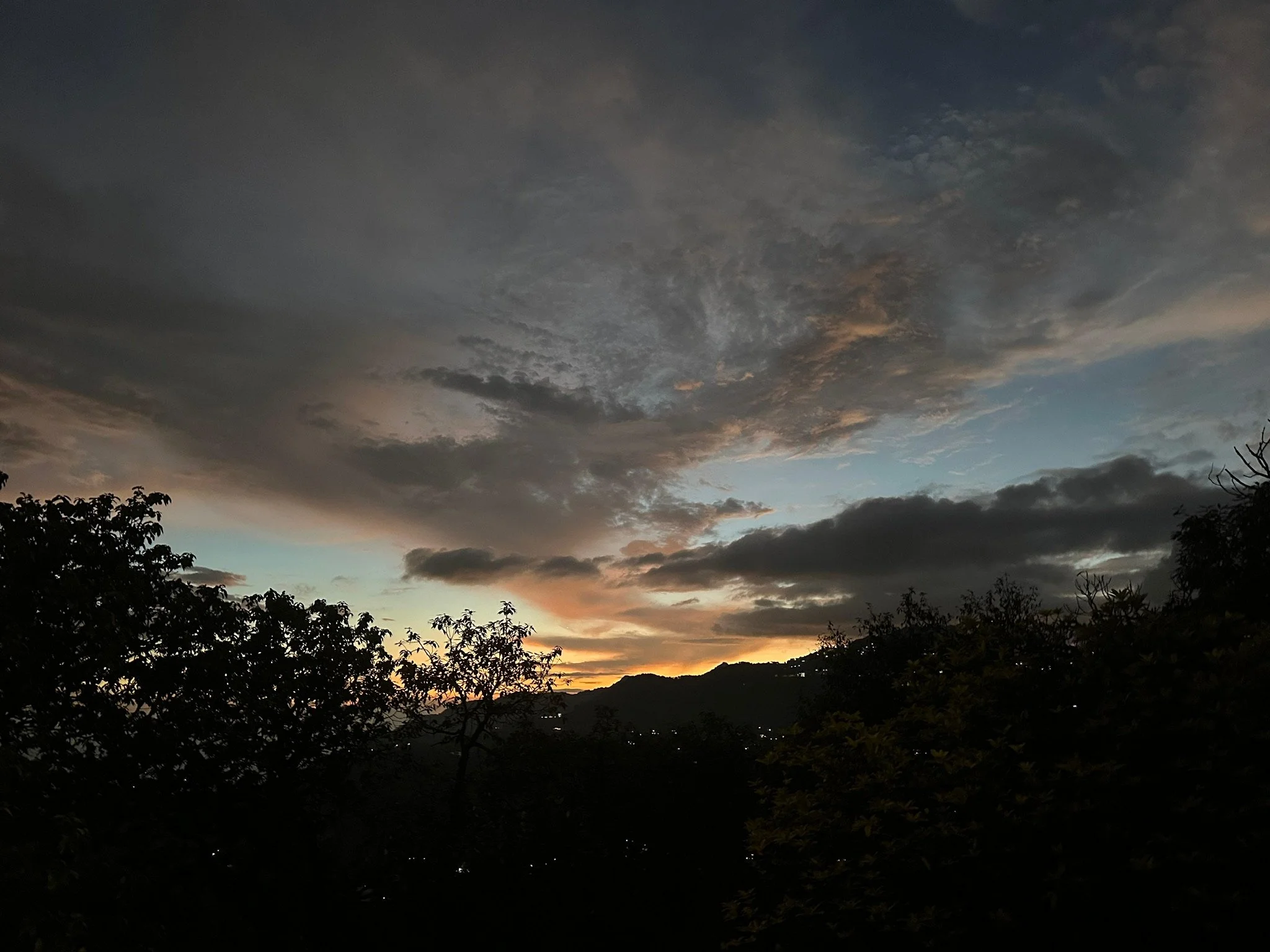 Sunset sky with dark clouds and a few patches of blue, overlooking silhouette of trees and mountains