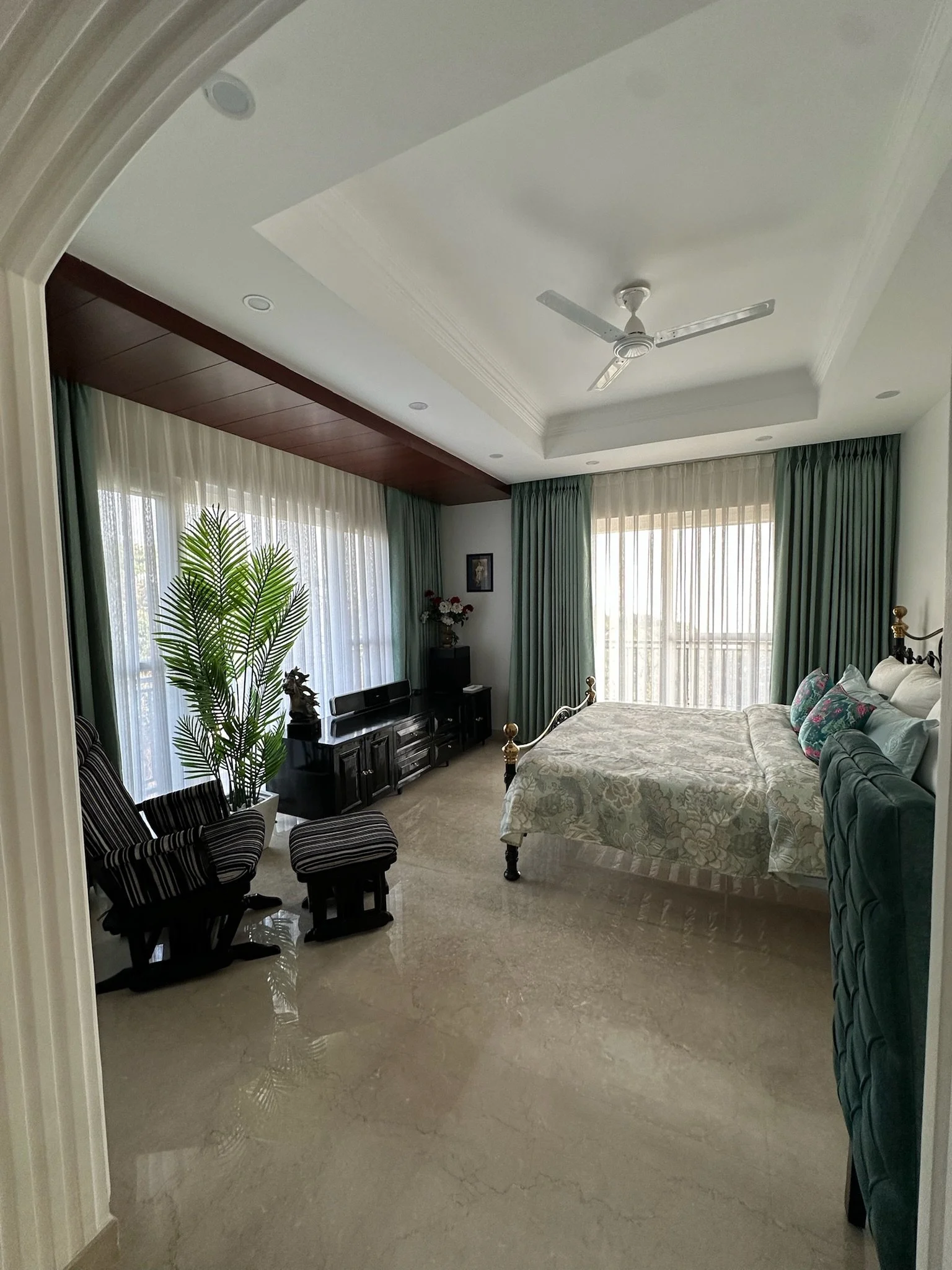A bedroom with a large metallic bed with ornate headboard, decorated with beige and red pillows, and a red blanket at the foot. Two dark wood nightstands on each side of the bed, a mirror on the wall, and two chairs with armrests and red floral pillows.