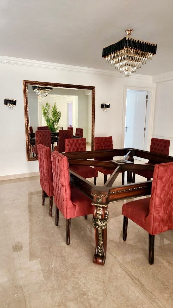 A dining room with a table surrounded by red velvet upholstered chairs, a large mirror on the wall reflecting a chandelier and a plant, and a chandelier hanging from the ceiling.