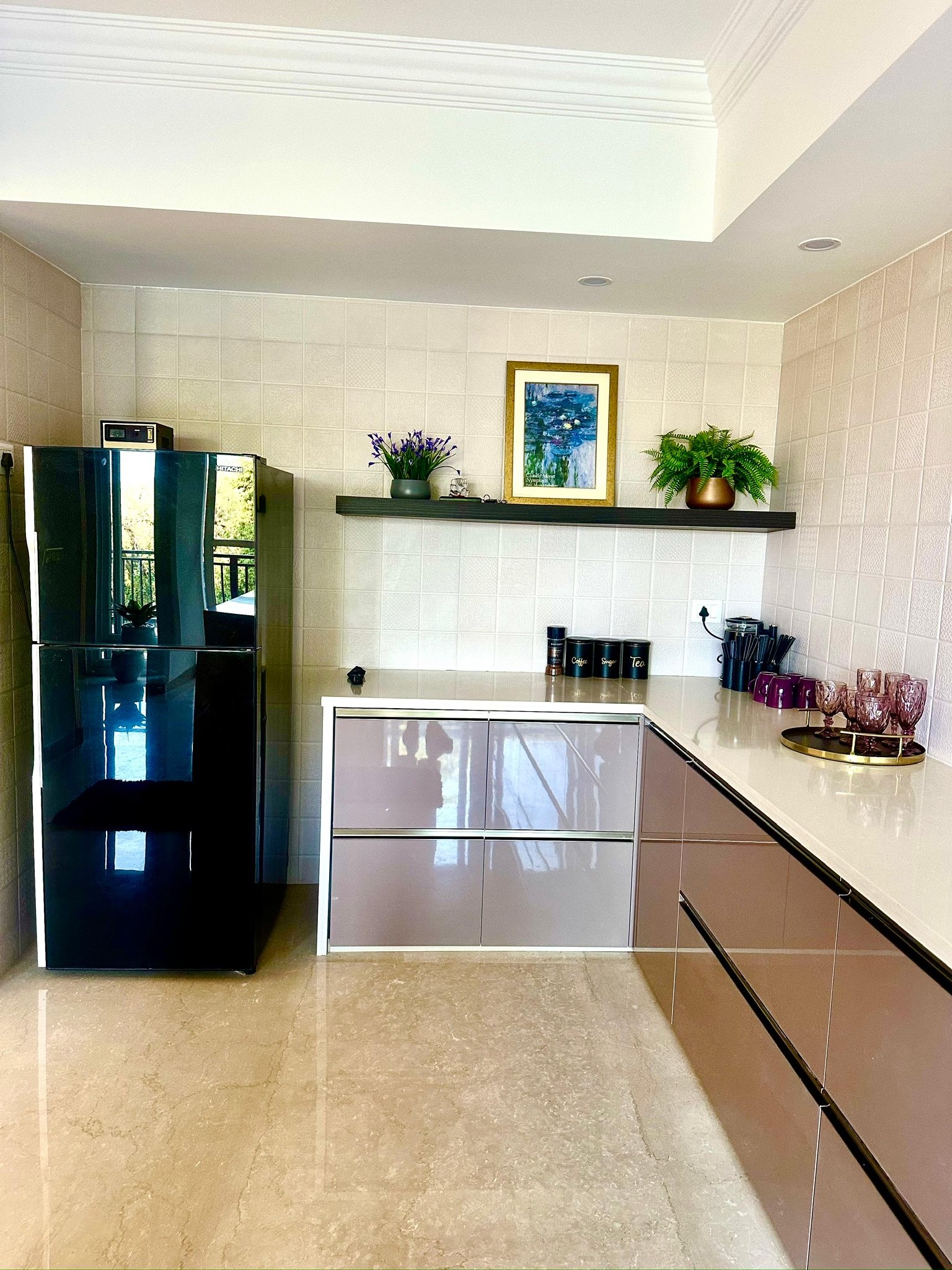 Modern kitchen with beige tiled walls and a shiny black refrigerator on the left. A white countertop with beige and purple cabinetry below. A black shelf with plants and art on top, and various cups and jars on the counter.