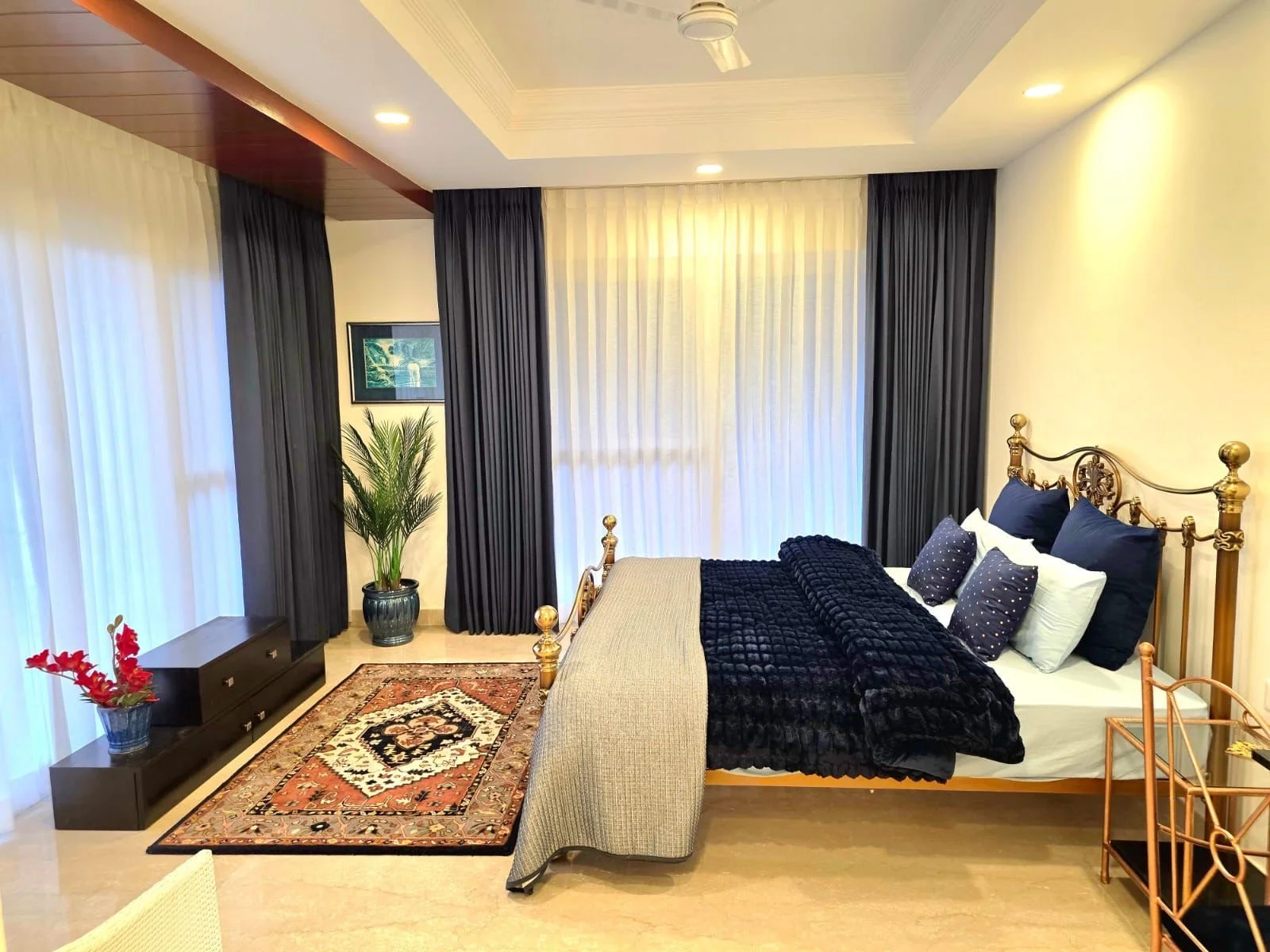 A bedroom with a gold metal bed frame, dark blue pillows and a black textured blanket, beige walls, sheer white and dark curtains, a plant in a black pot, a picture on the wall, a rug with a traditional pattern, a black console table, and a rose gold cart.