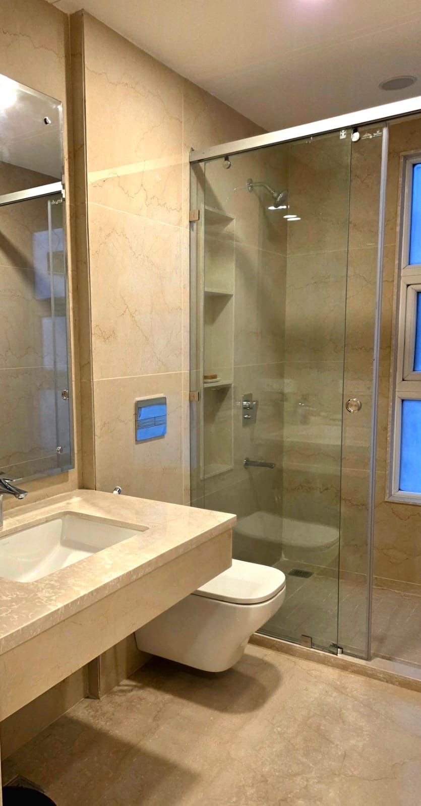 A modern bathroom with a beige marble vanity, a rectangular sink, a wall-mounted toilet, a glass-enclosed shower with built-in shelves, a window with frosted glass, and beige tiled walls.