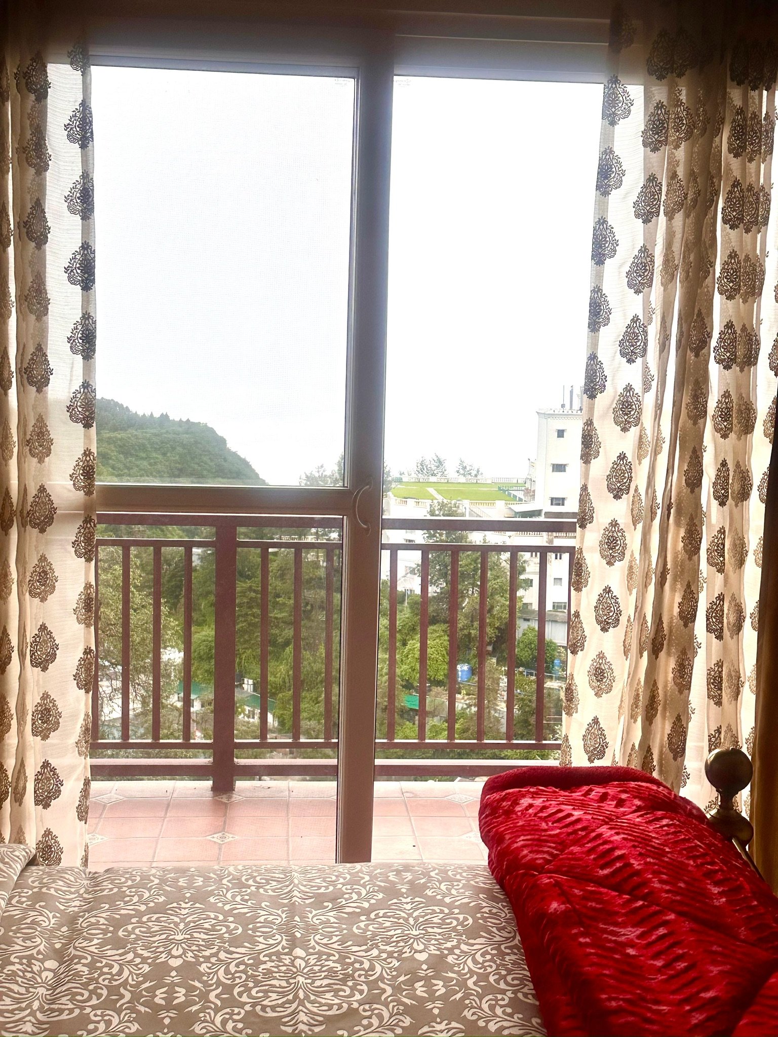 View from inside a bedroom showing a large window with patterned curtains, a balcony railing, trees, and buildings outside. A bed with patterned bedding and a large red pillow is in the foreground.