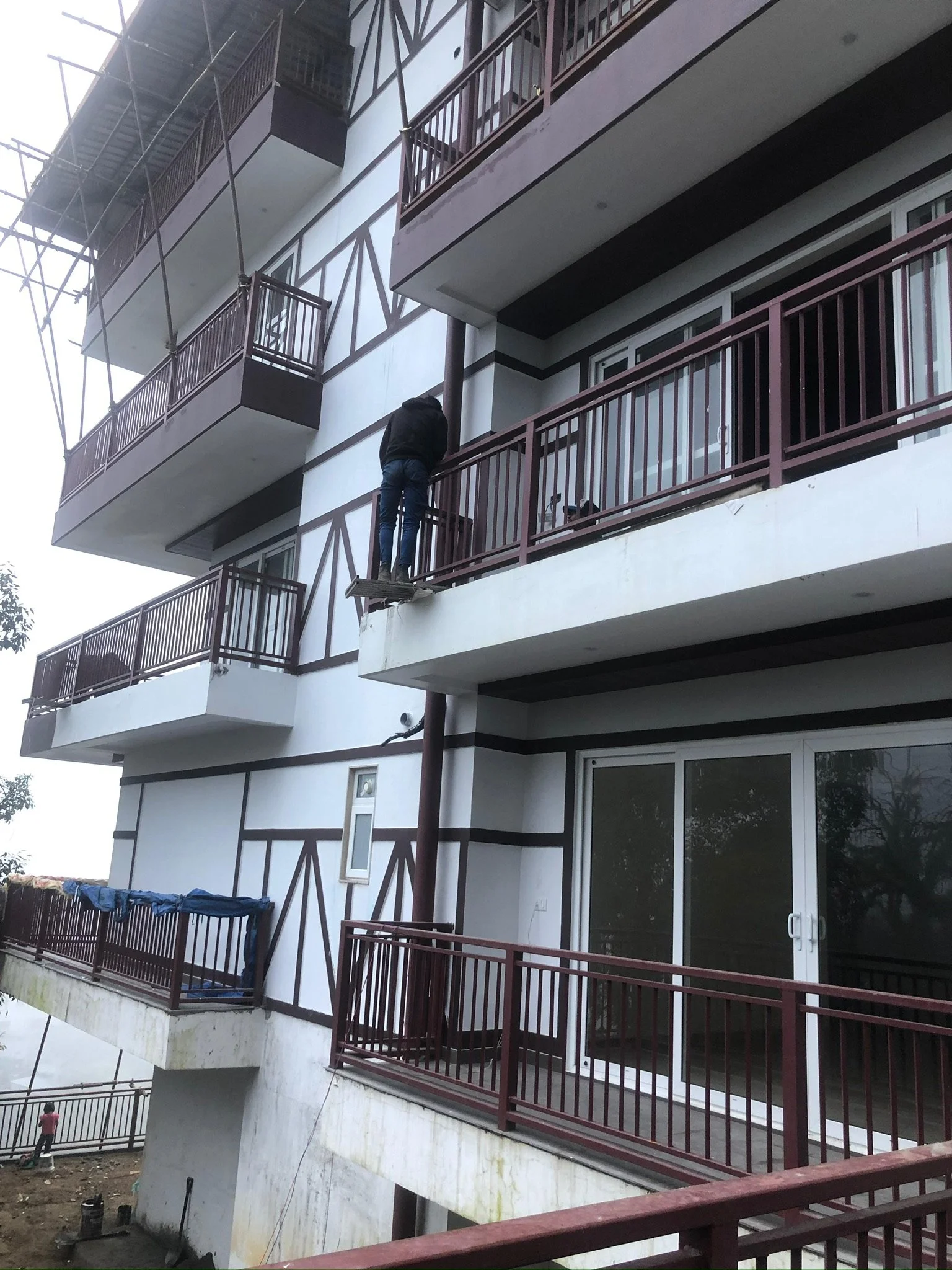 Construction worker standing on a small wooden platform, working on the balcony railing of a multi-story building with brown railings and white walls.