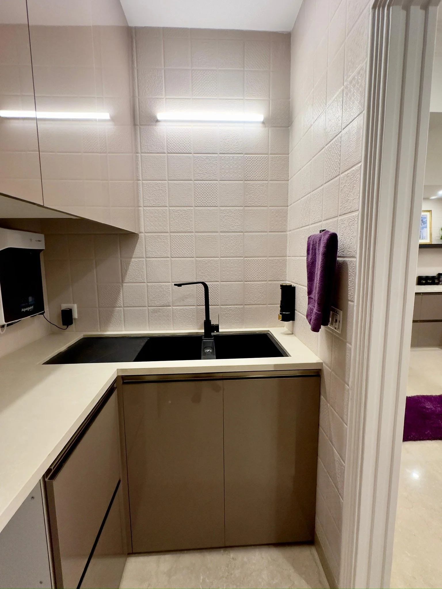 Small kitchenette with black sink, beige cabinets, purple towel, wall-mounted soap dispenser, and bright overhead lighting.