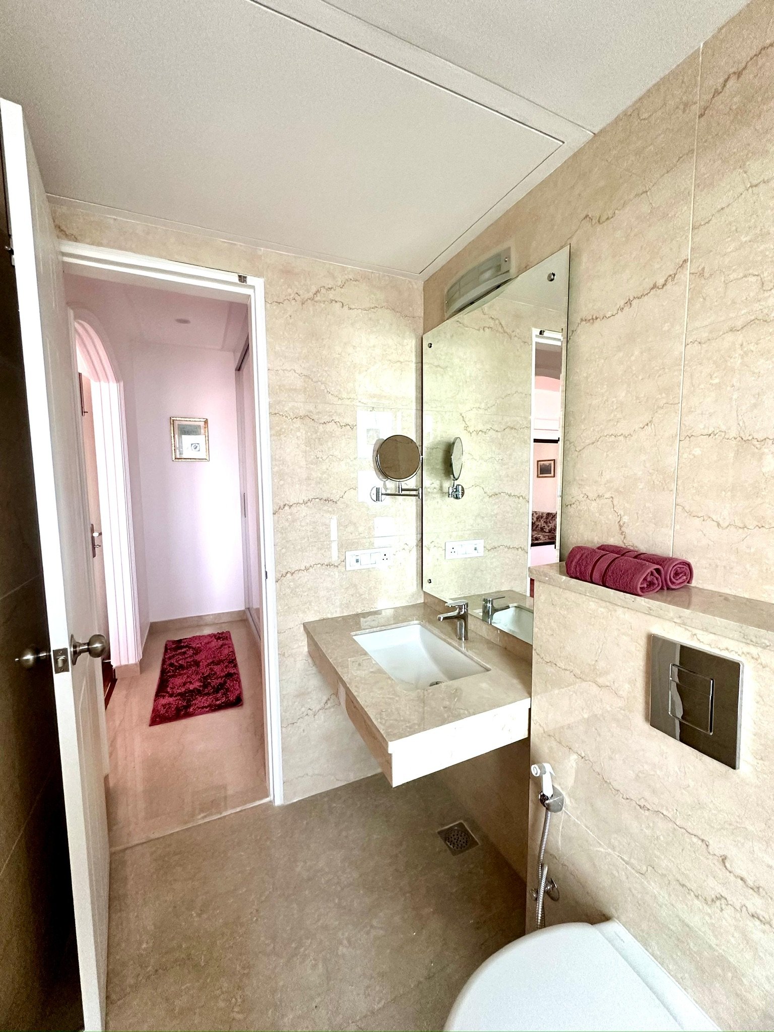 A bathroom with beige marble walls, a small sink with a marble countertop, and a large mirror. There are pink towels on a ledge and a door leading to a hallway with pink walls and a red rug.