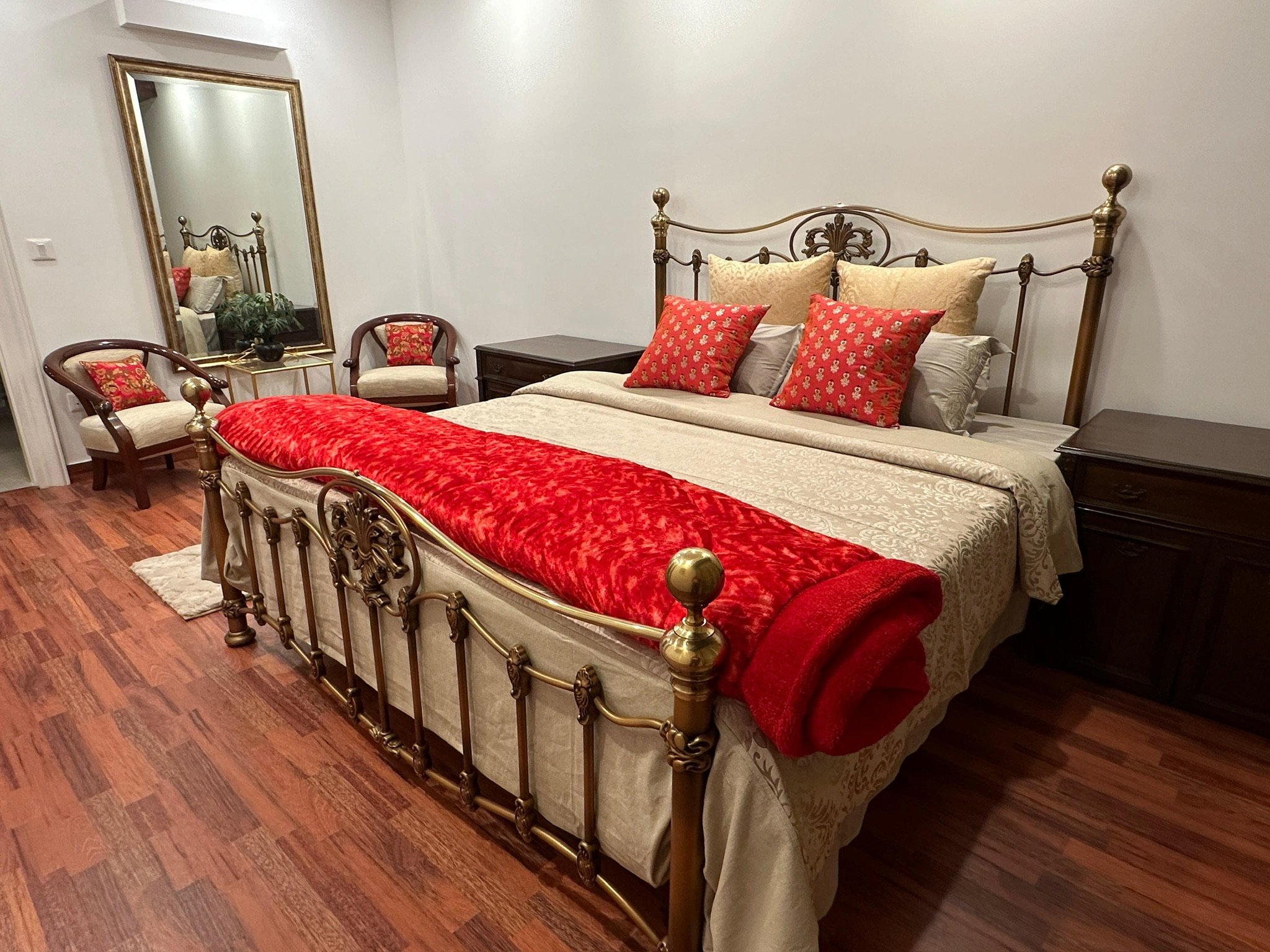 A bedroom with a large metallic bed with ornate headboard, decorated with beige and red pillows, and a red blanket at the foot. Two dark wood nightstands on each side of the bed, a mirror on the wall, and two chairs with armrests and red floral pillows.