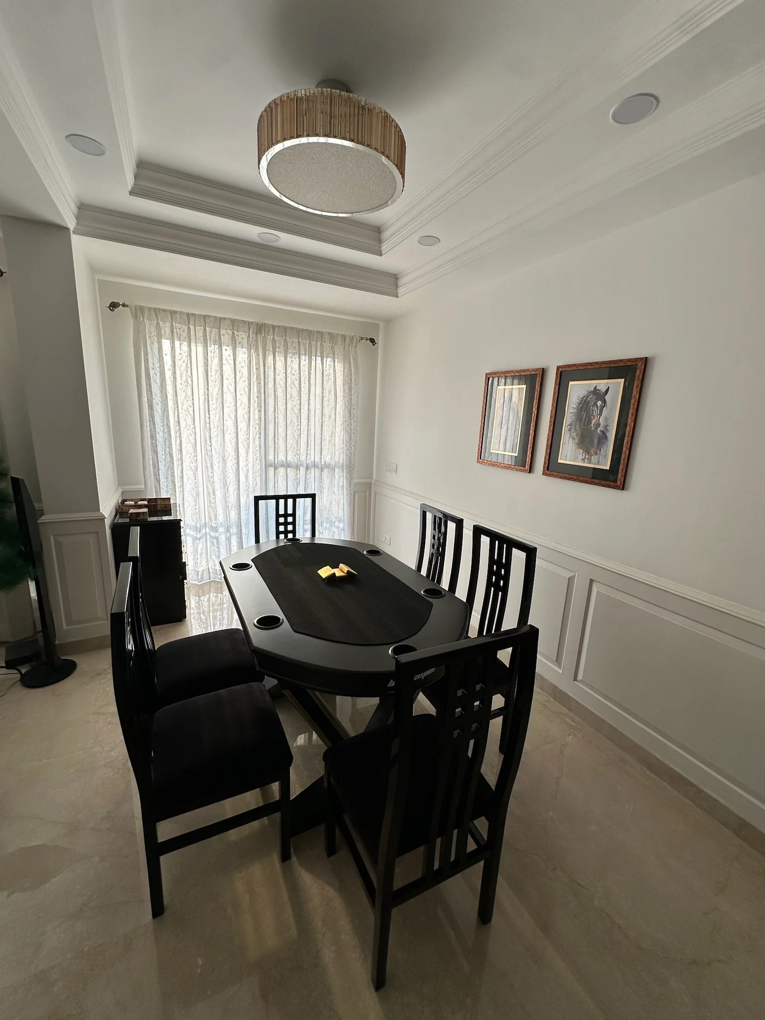 Dining room with six black chairs around a black oval table, two framed horse paintings on the wall, elegant chandelier overhead, and light-colored walls and flooring.