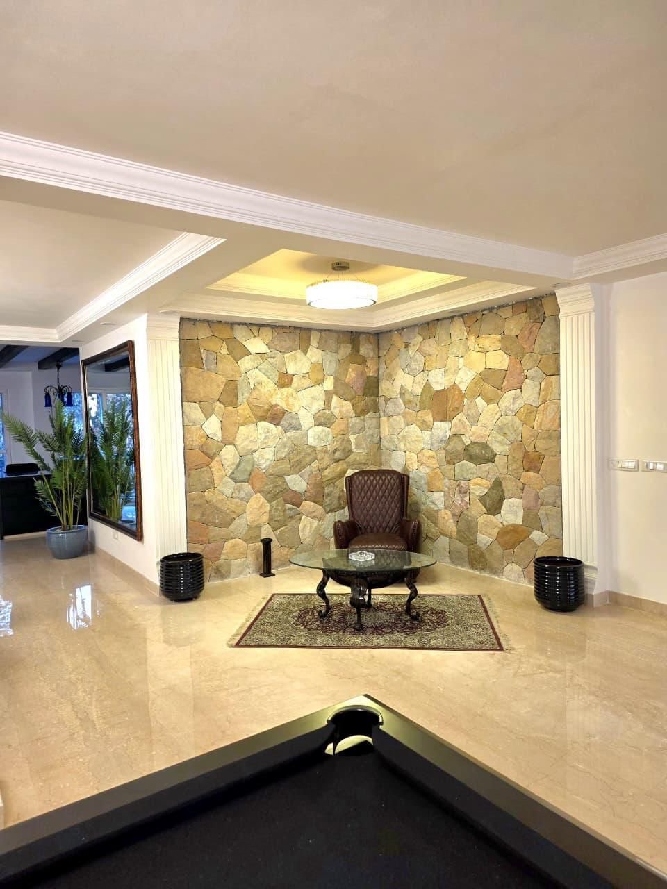 Interior of a living room with a stone accent wall, a brown armchair, glass coffee table, small area rug, and two black heater vents on either side of the chair.