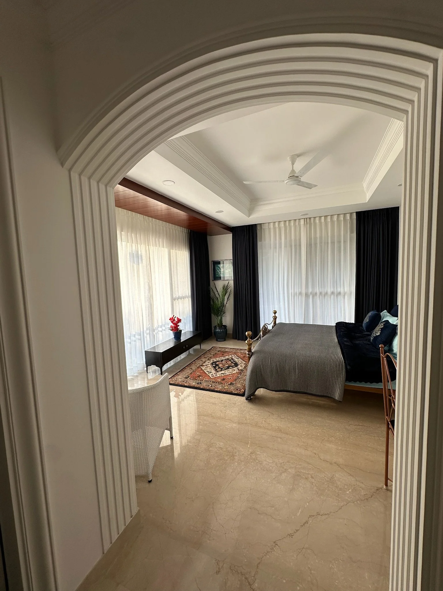 A bedroom with a metal bed frame, cream and burgundy pillows, a plaid blanket, a Persian rug, beige curtains, and a balcony door with blue sky outside.