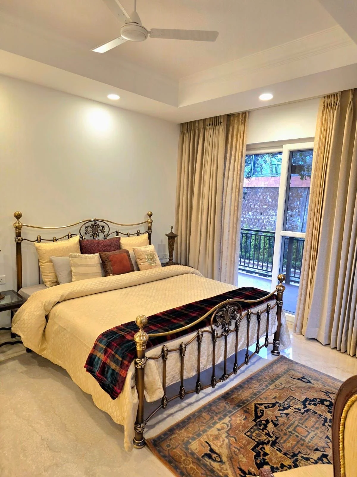 A bedroom with a metal bed frame, cream and burgundy pillows, a plaid blanket, a Persian rug, beige curtains, and a balcony door with blue sky outside.