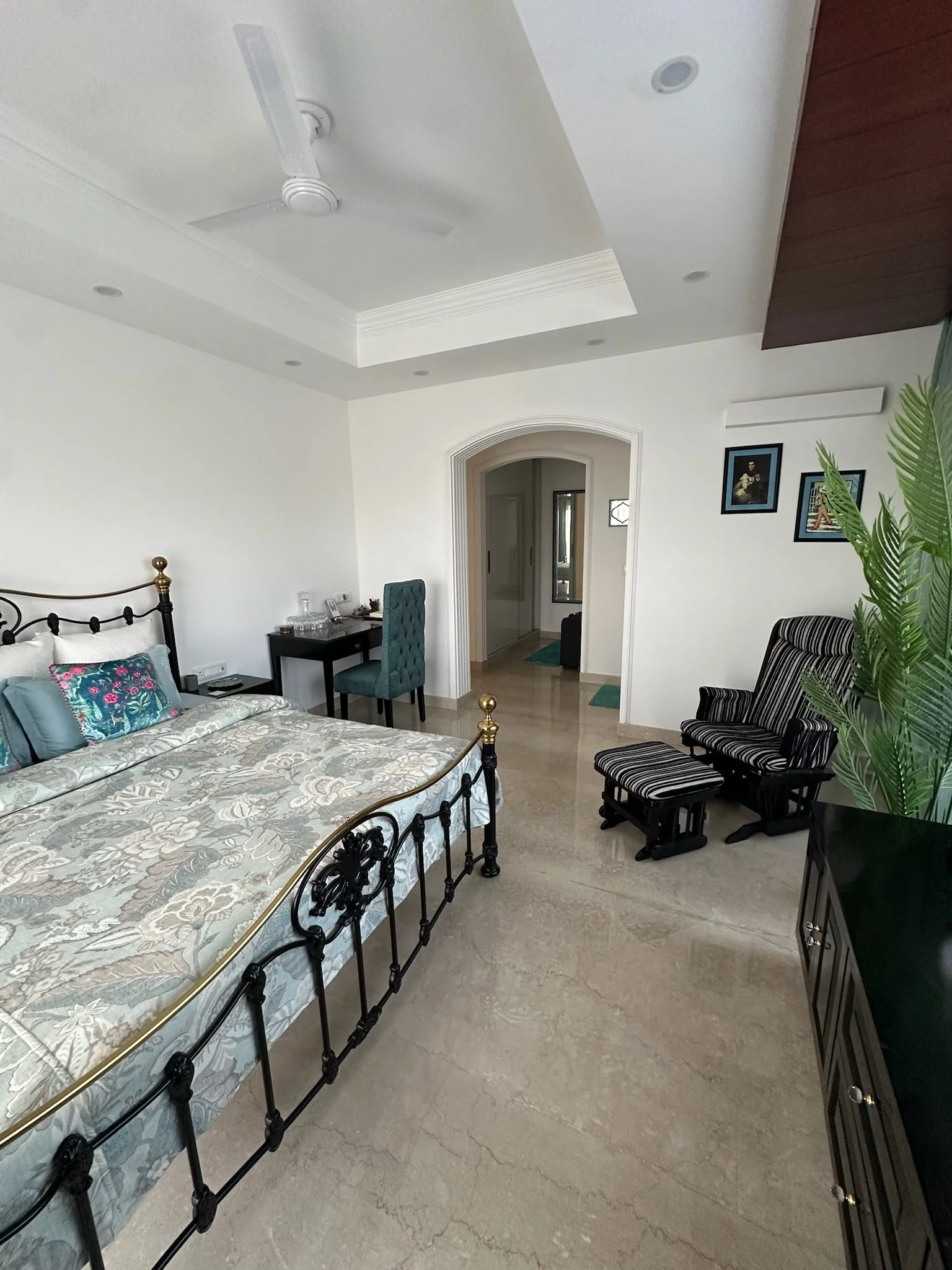 A cozy corner with two wooden armchairs with beige cushions and red decorative pillows, a gold metal side table with a black vase holding a large green plant, a tall mirror on the wall, a beige rug on a hardwood floor, and a glimpse of a bed with a brass headboard reflected in the mirror.