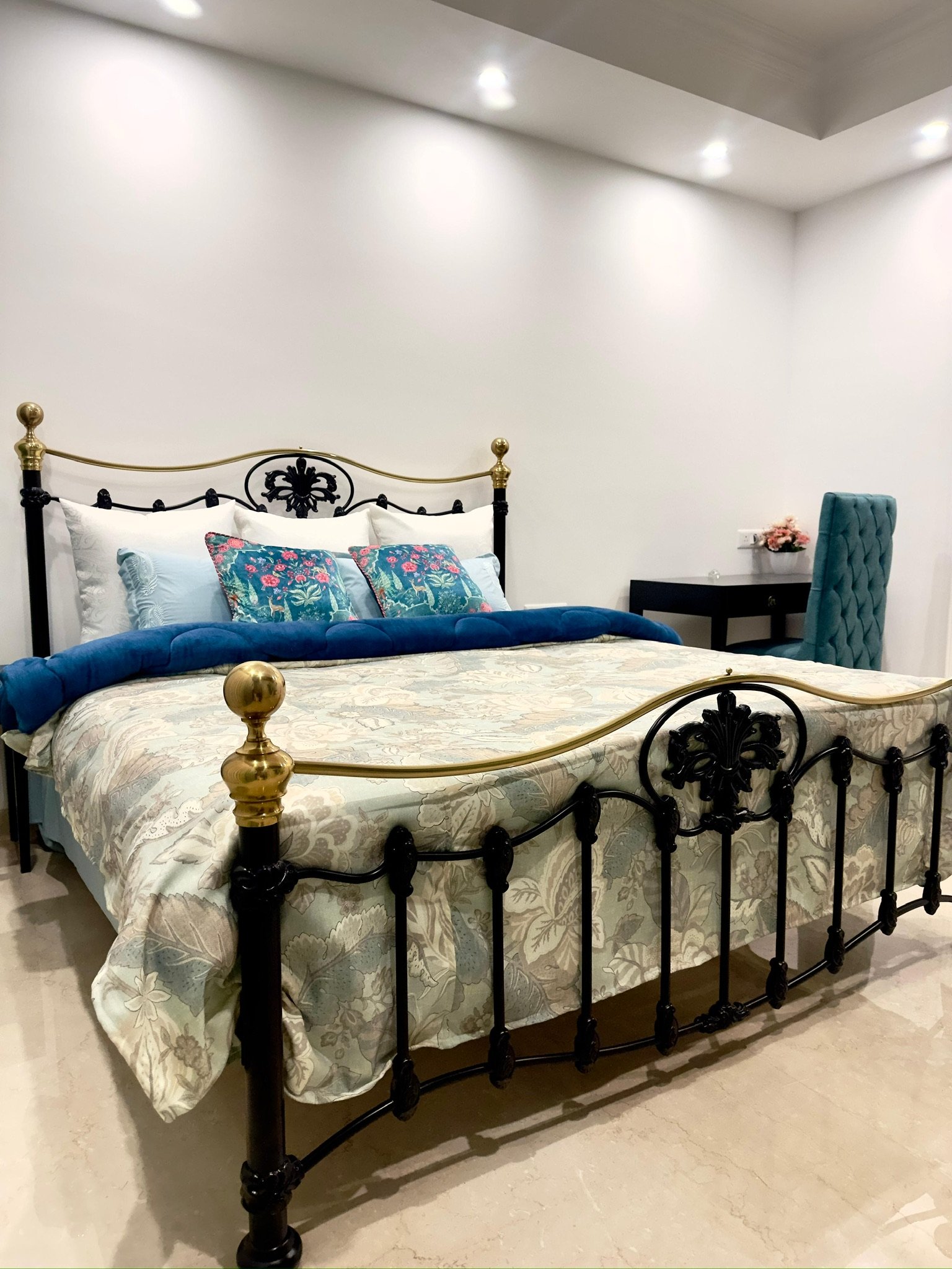 A bedroom with a black and gold ornate metal bed frame, white bedding with a floral pattern, blue pillows, and a padded turquoise chair next to a small black desk with a pink flower arrangement.