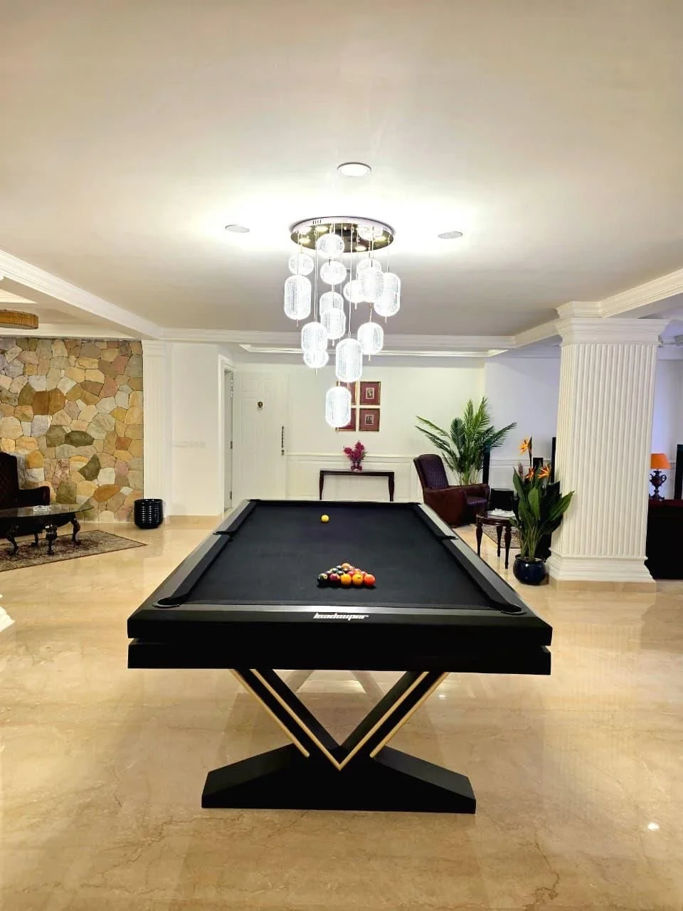 Indoor lounge with a black pool table set up for billiards, positioned under a modern chandelier with multiple hanging glass lights, surrounded by armchairs, plants, and decorative wall art.