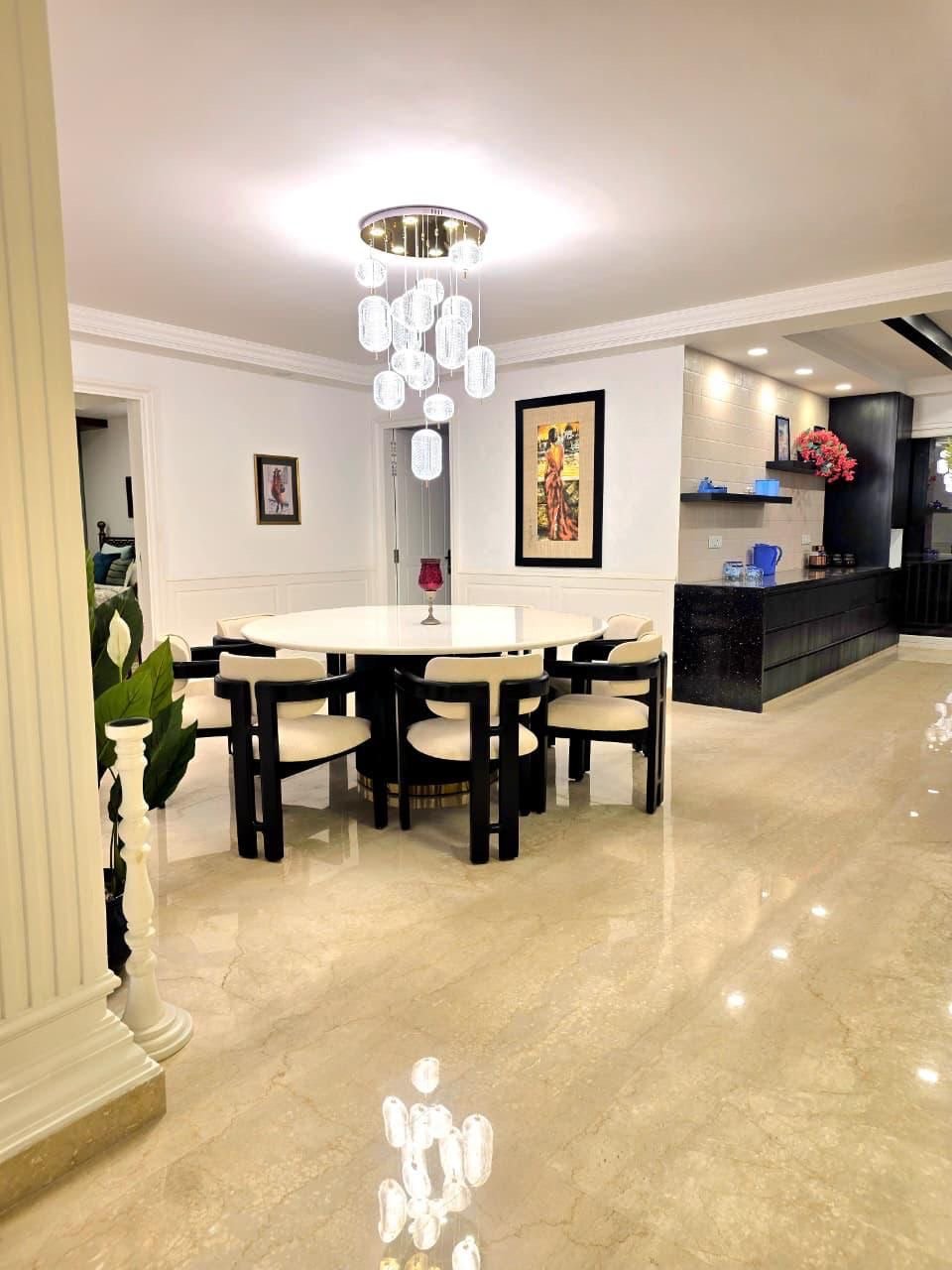 Modern dining room with a white round table, eight beige chairs with black frames, a chandelier with hanging glass orbs, framed artwork on white walls, beige marble flooring, a black kitchen counter with shelves, and a doorway leading to another room.