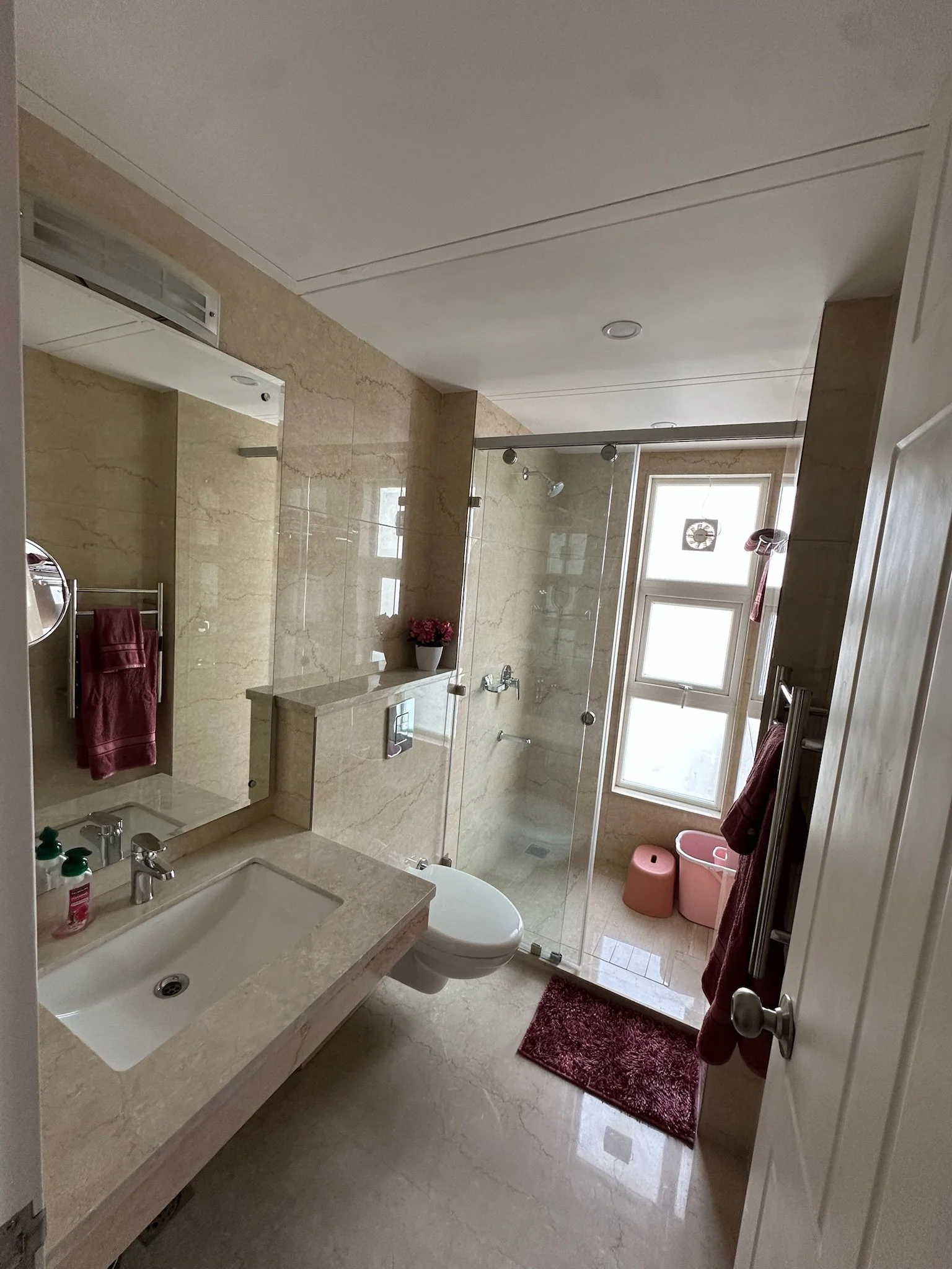 A small bathroom with beige marble walls and floor, featuring a sink with a beige countertop, a large mirror, a small shelf with a black mirror, a towel rack with turquoise towels, a vibrant pink flower arrangement on the wall, and a teal rug on the floor.