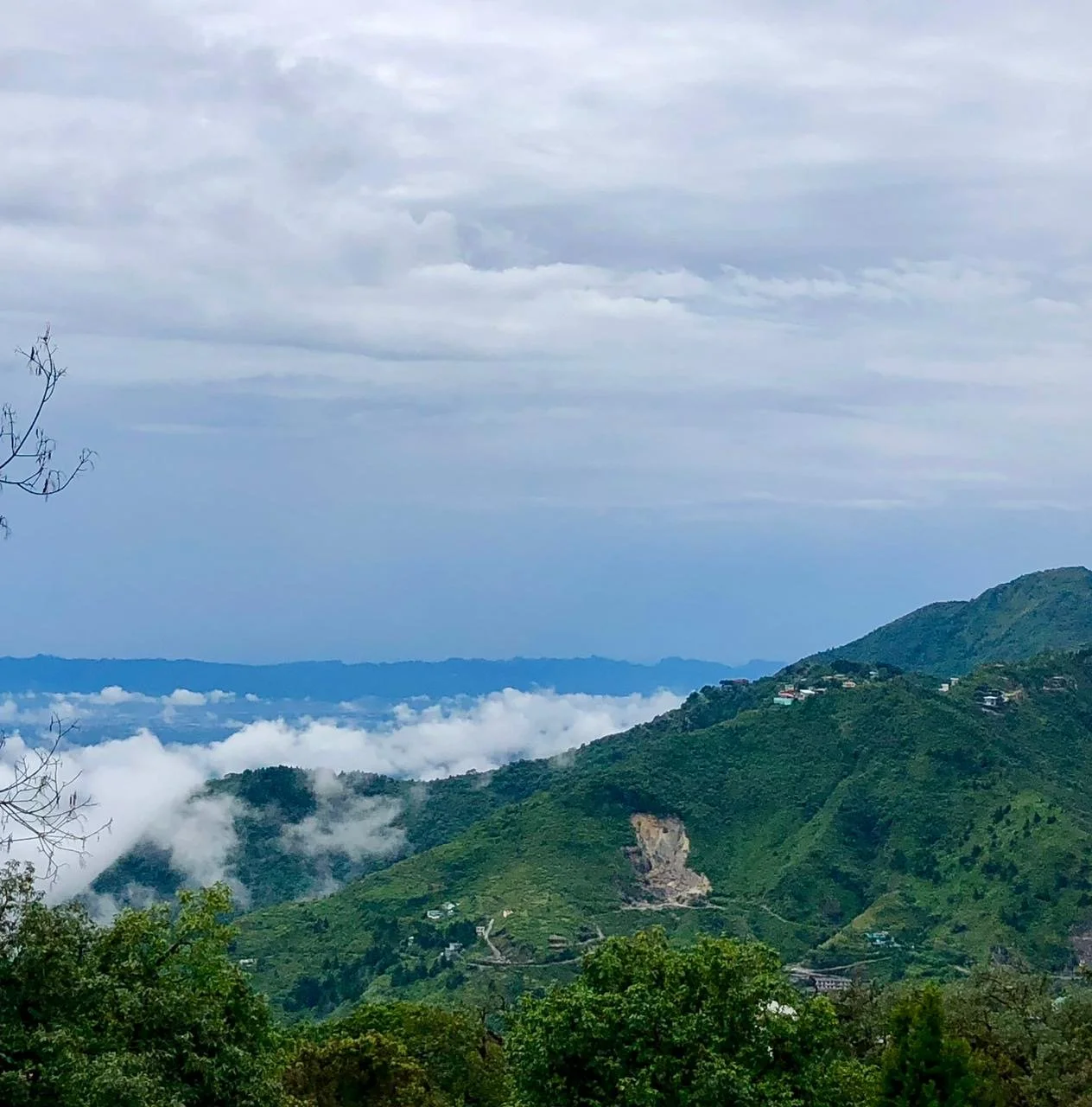 Mountain landscape with green hills, scattered houses, and fog in the valleys under a cloudy sky.