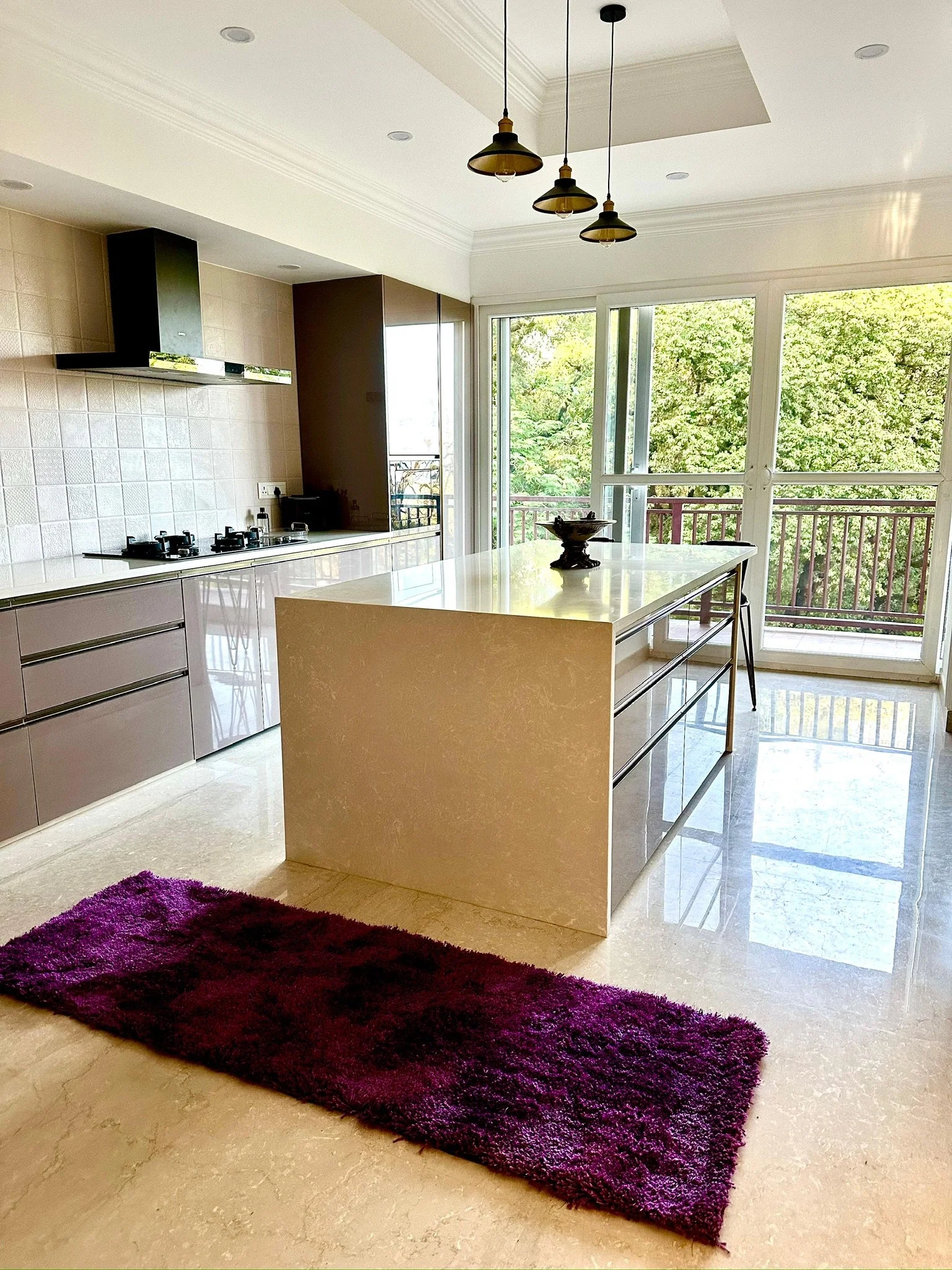 Modern kitchen with a beige marble island, purple rug, and large glass sliding door leading to a balcony with green trees outside.