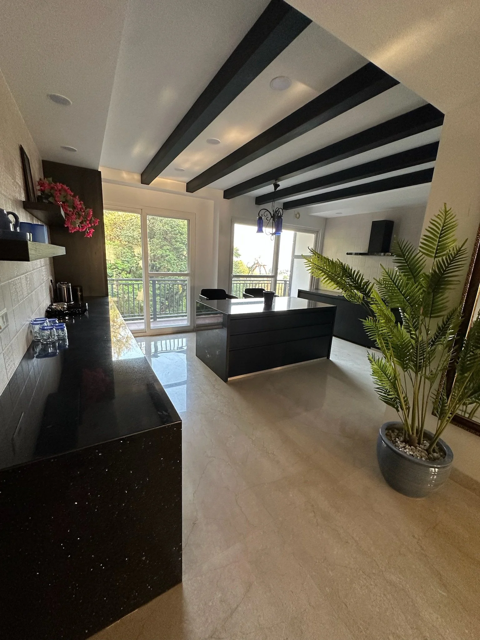 Modern dining room with black ceiling beams, a black dining table with chairs, a chandelier with blue lights, large windows with a balcony outside, and a potted plant.