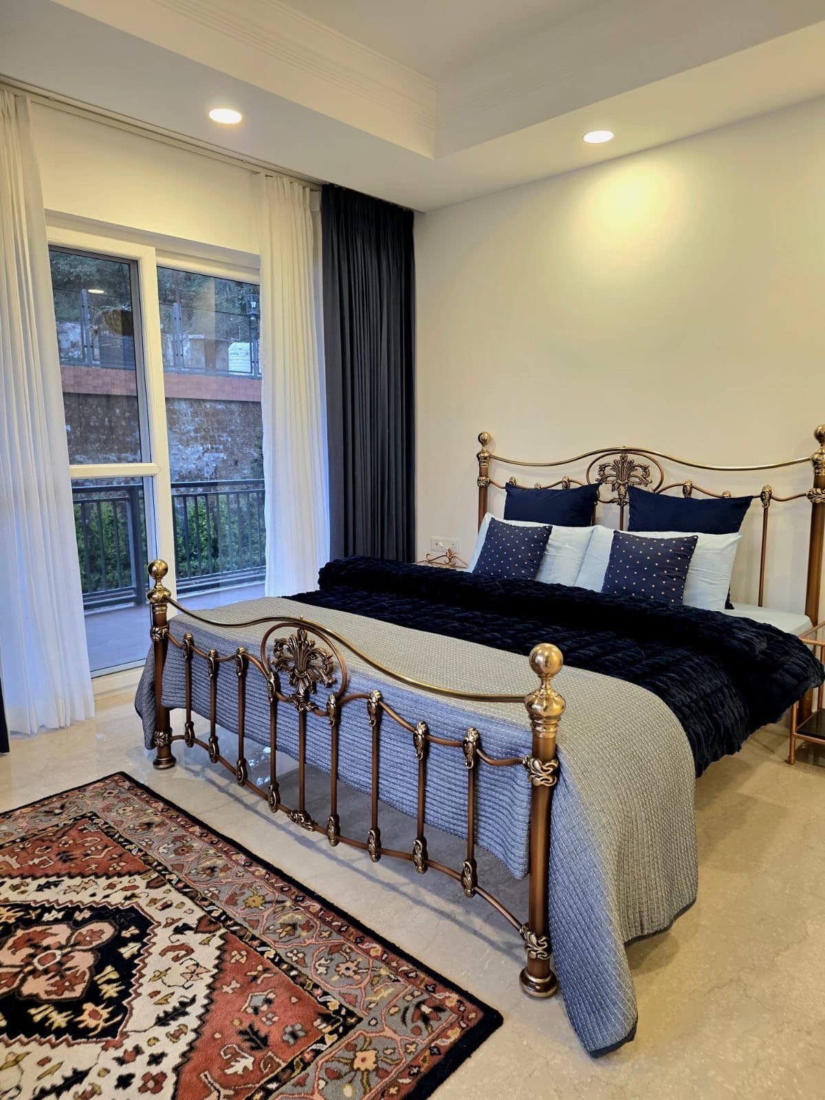 A bedroom with a brass bed frame, blue and white bedding, a patterned area rug, and a sliding glass door with white and dark curtains leading to a balcony.