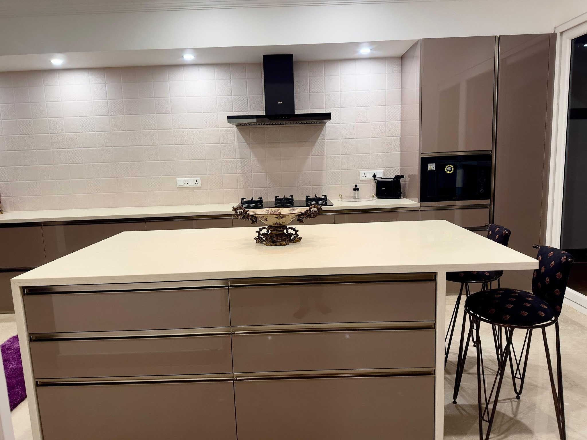 Modern kitchen with beige cabinets, a white kitchen island, black bar stools, a tiled backsplash, a stovetop, a faucet, and small appliances.