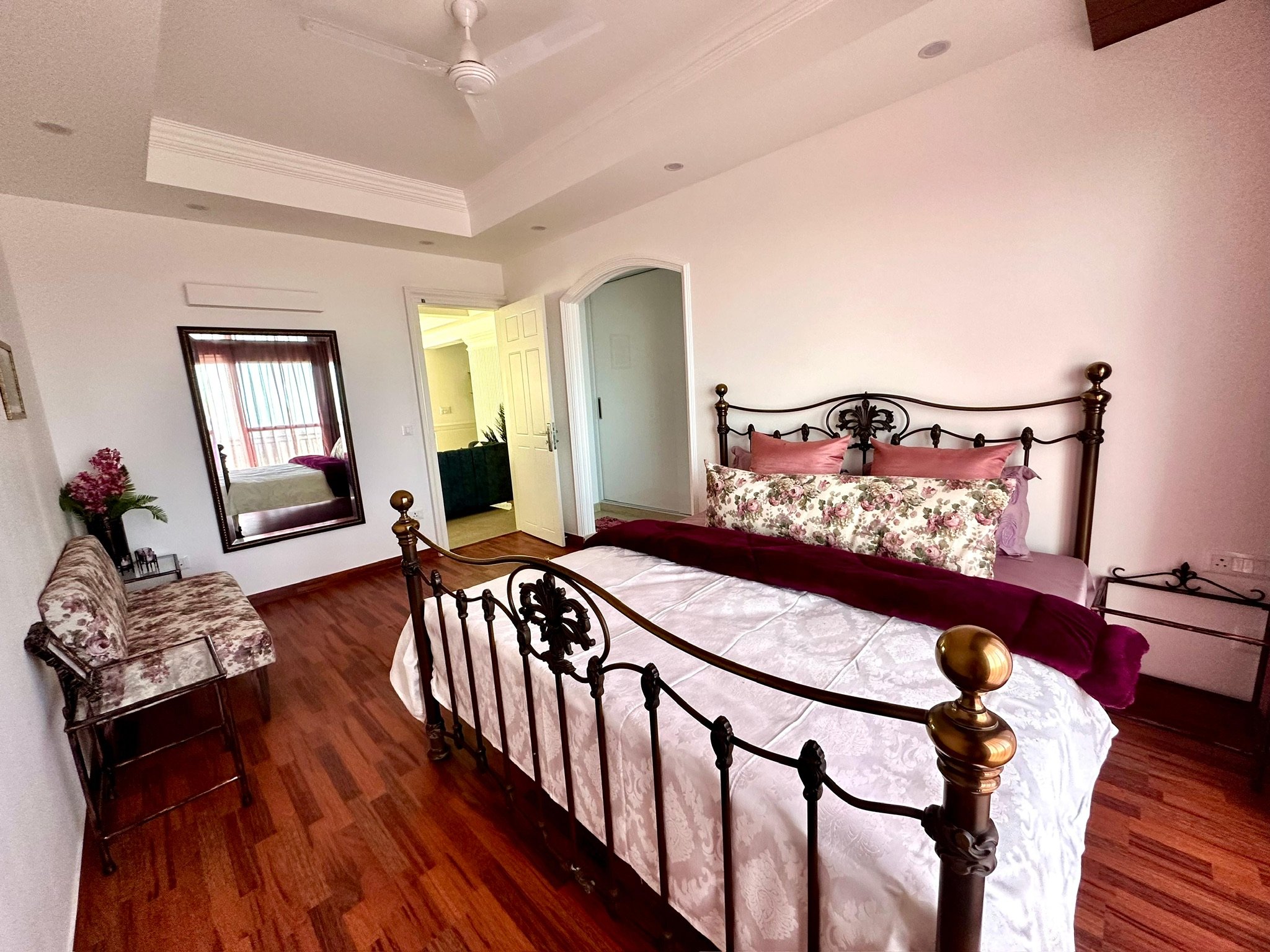 A bedroom with a four-poster bed, floral pillows and duvet, a vintage style bench with floral upholstery, a mirror on the wall, a small table with flowers, and a door leading to another room with green furniture. The room has wooden flooring and a ceiling fan.
