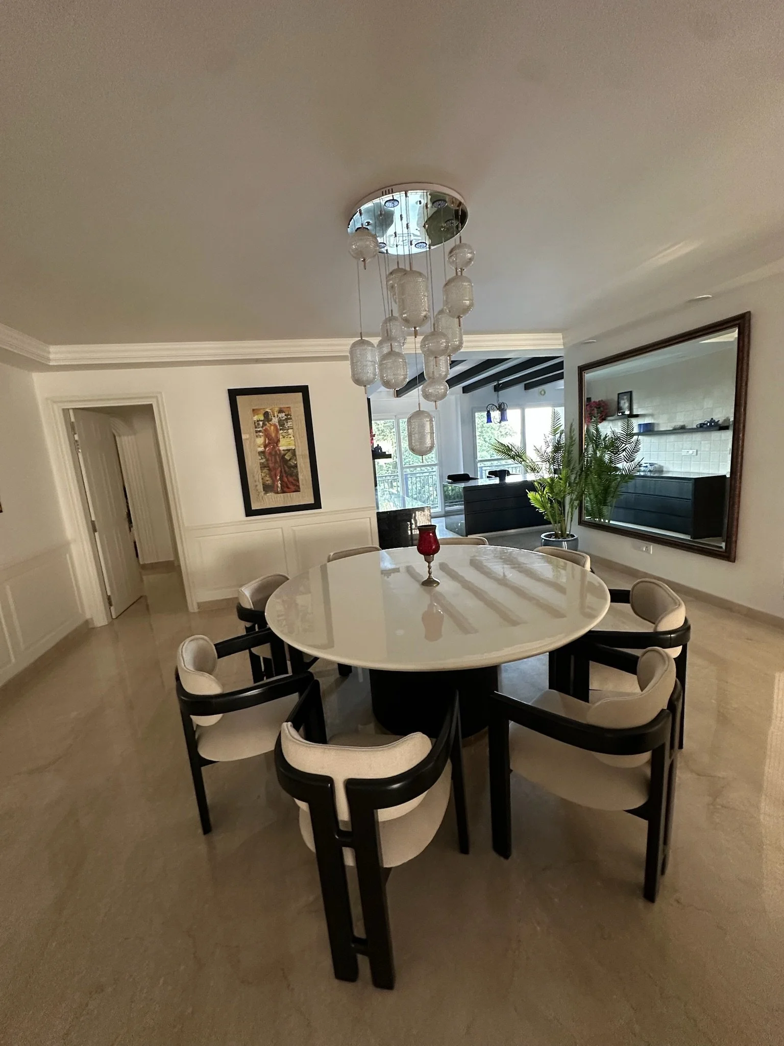 A dining room with a round table, ten chairs, a chandelier, large wall mirror, plants, a stone accent wall, and a small seating area with an armchair.