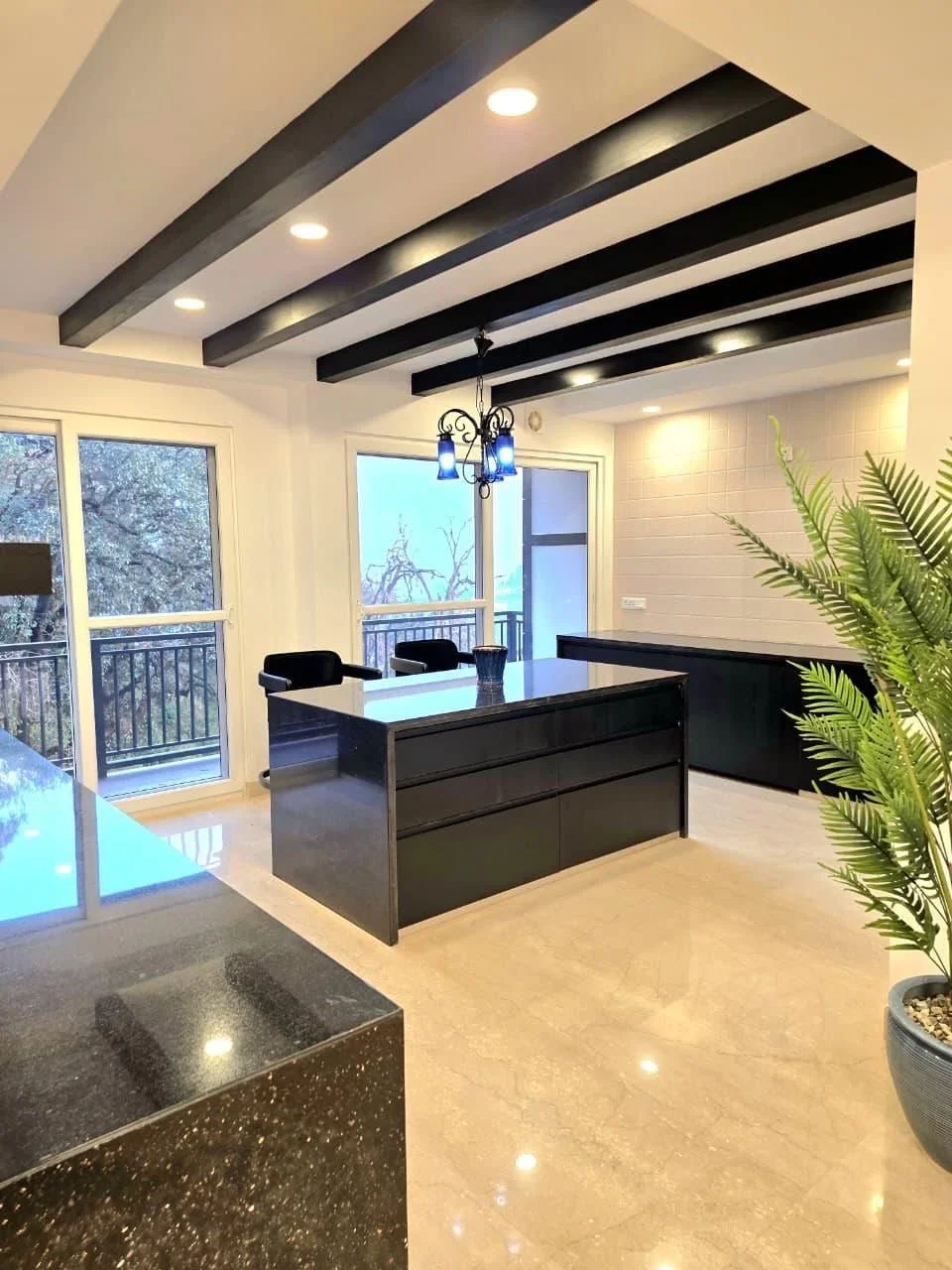 Modern dining room with black ceiling beams, a black dining table with chairs, a chandelier with blue lights, large windows with a balcony outside, and a potted plant.