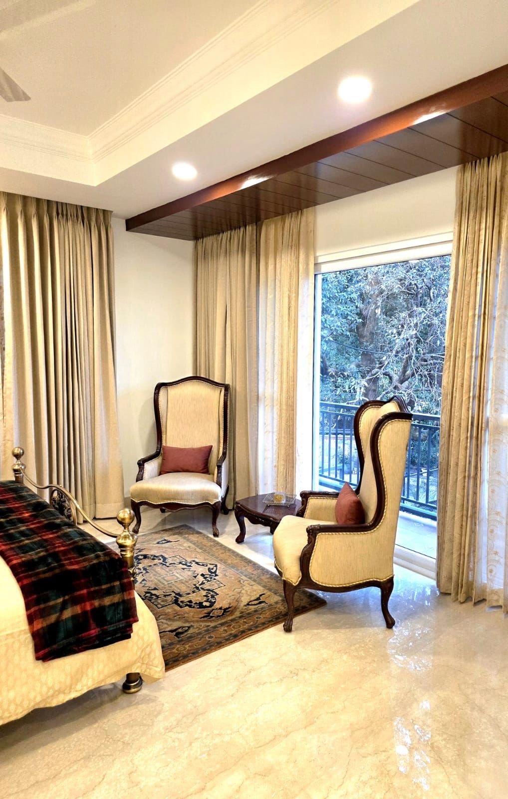Living room with two beige armchairs with wooden frames, small side table, Persian-style rug, large window with cream curtains, outside snowy trees, marble floor, ceiling with recessed lighting.