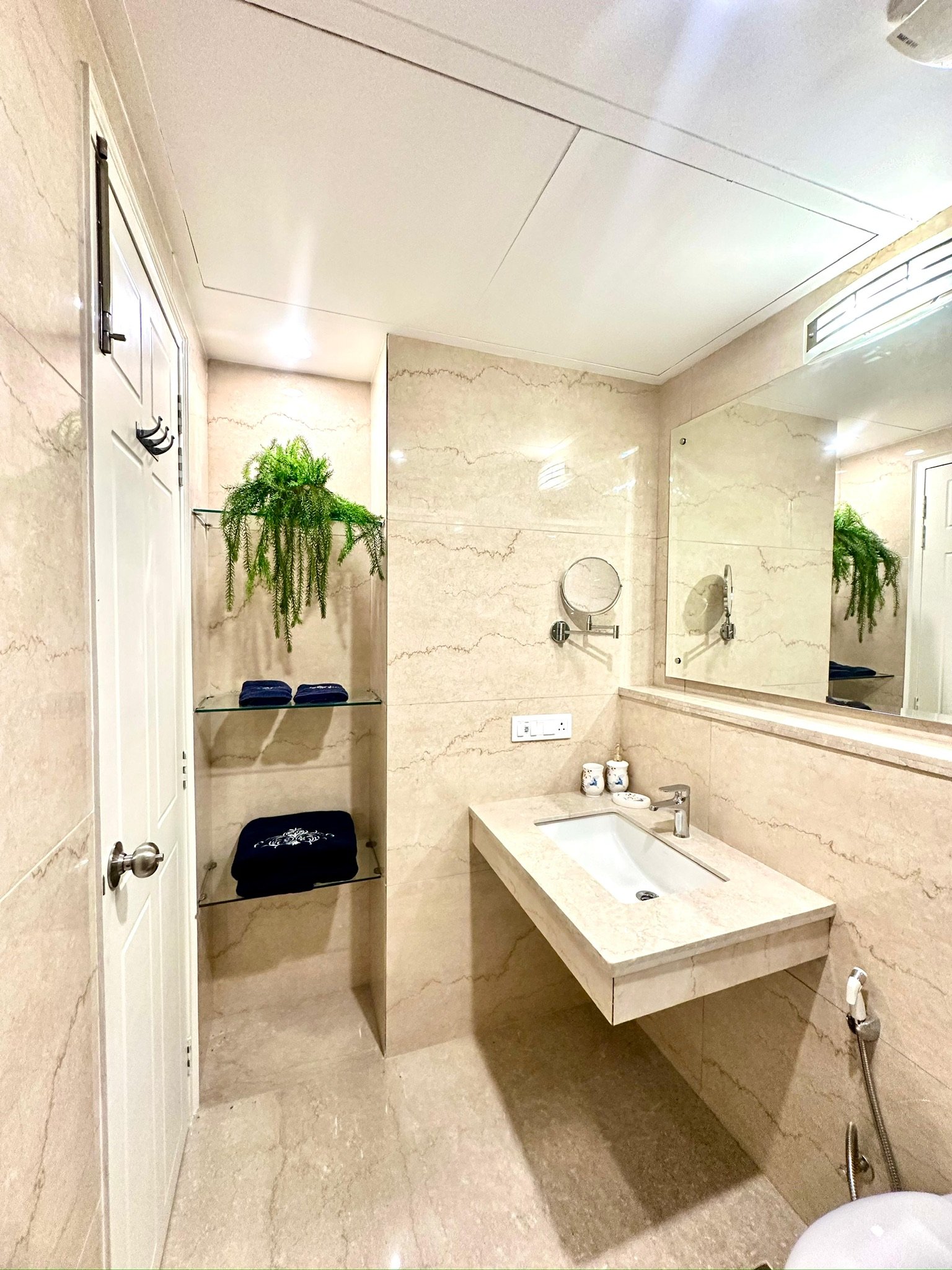 A modern bathroom with beige marble walls and flooring, a rectangular sink with a marble countertop, a large mirror, a wall-mounted magnifying mirror, potted plant on glass shelves, and folded towels.