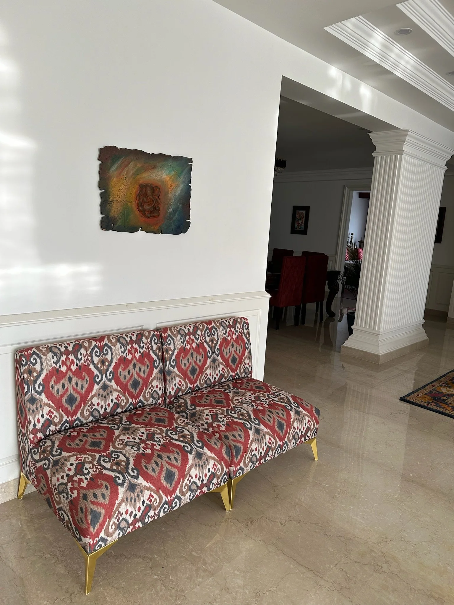 A spacious living room with white walls, a ceiling fan, a chandelier, two blue sofas with pillows, a wooden coffee table, a patterned rug, a tall green potted plant, and a framed painting on the wall.