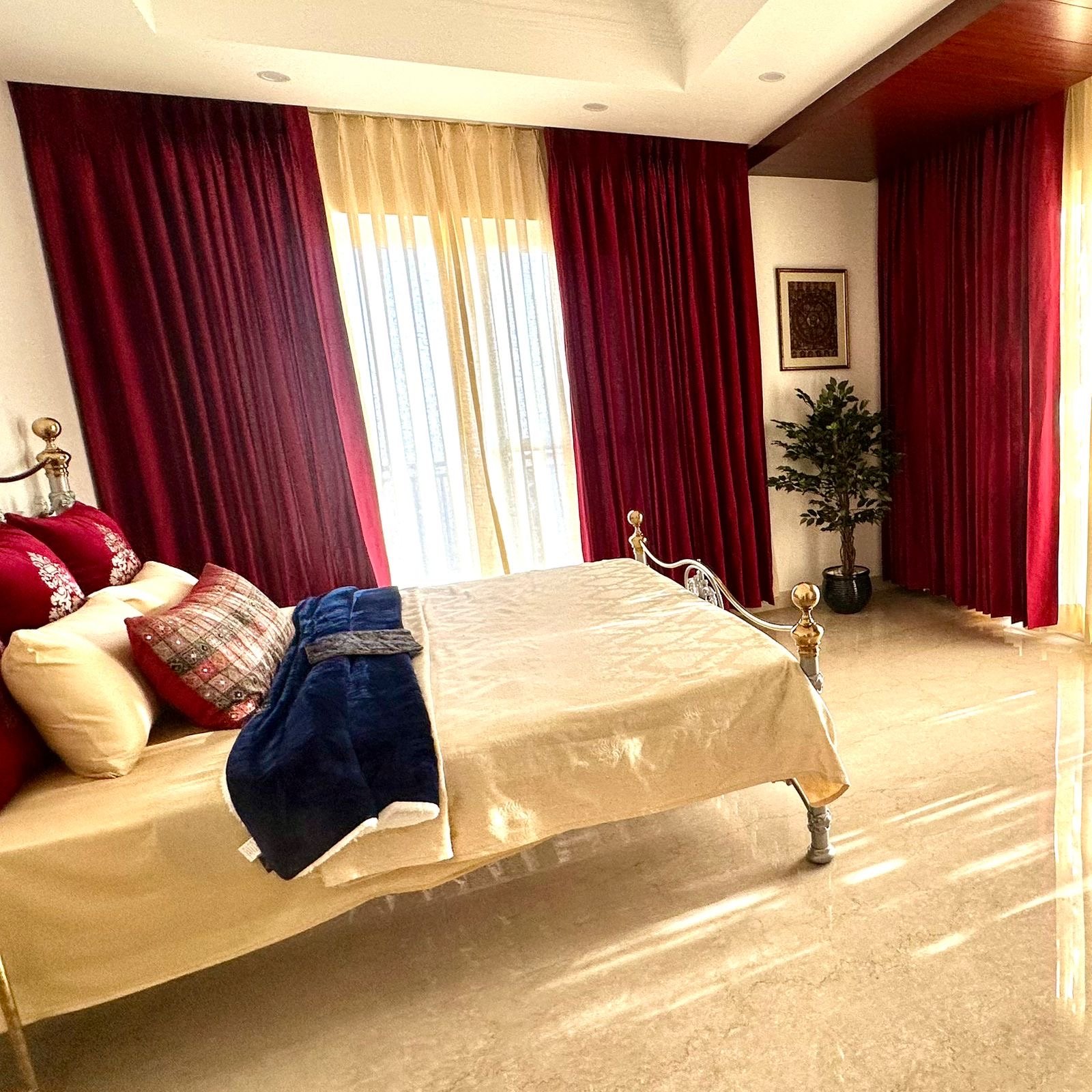 A bedroom with a bed, red and cream curtains, a framed picture, a potted plant, and a wooden floor.
