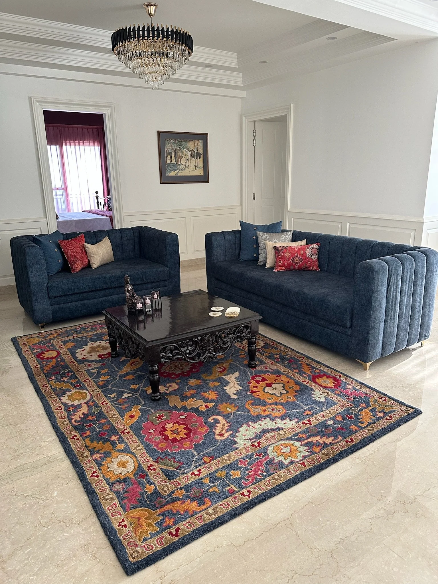 Elegant living room with a crystal chandelier hanging from a white ceiling, cream walls with crown molding, dark blue velvet sofas with pillows, and a place with a large green plant.