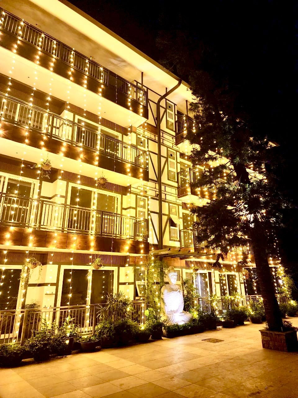 Multi-story building decorated with numerous yellow string lights, with potted plants and small statues near the entrance, and a large tree to the right.
